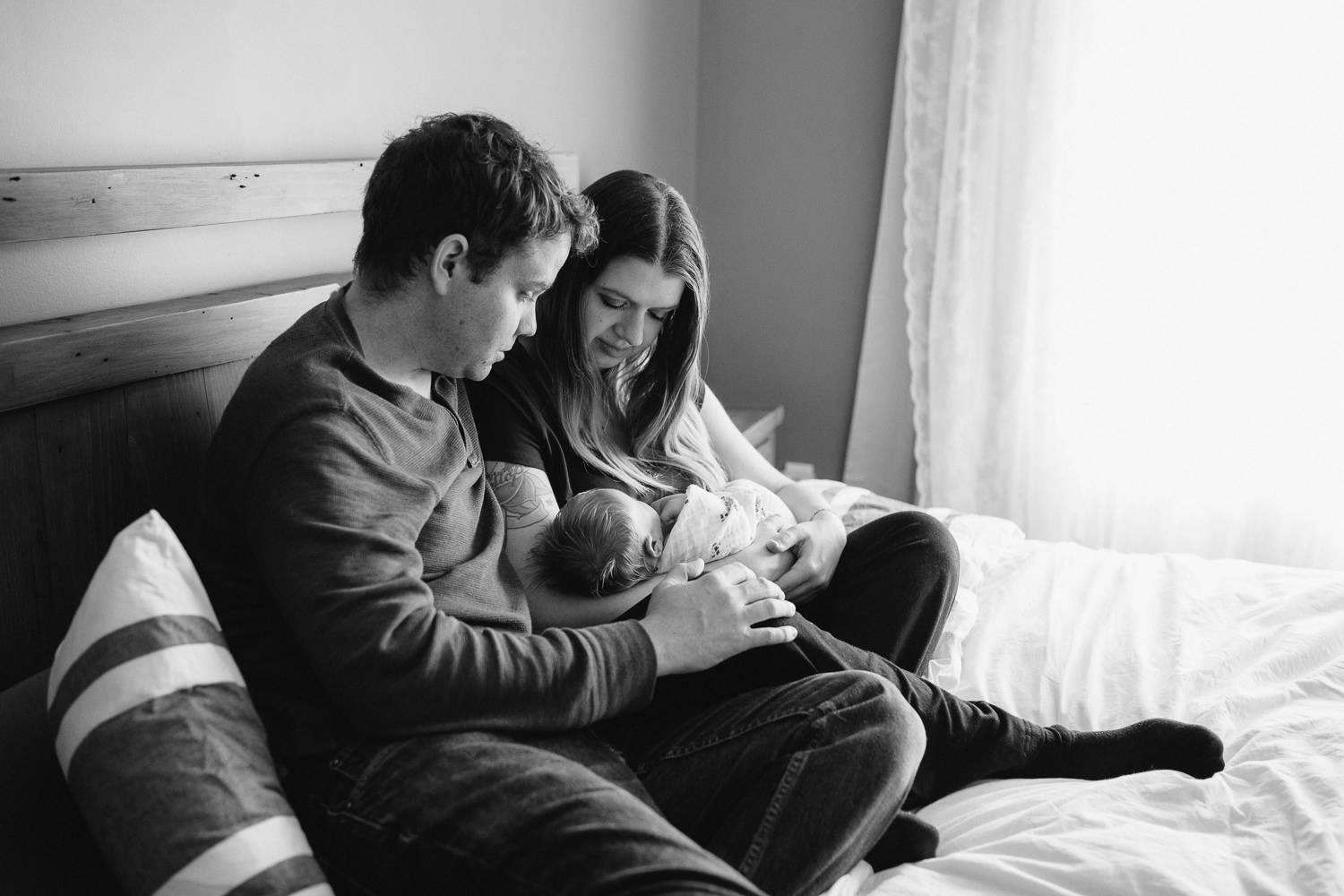 family of 3, new parents sitting on bed holding and looking at 2 week old baby girl with red hair - Stouffville Lifestyle Photography