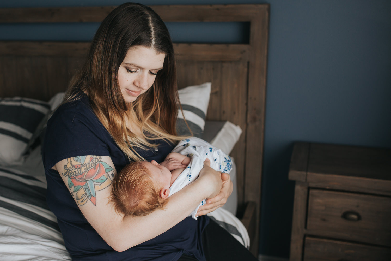 first time mom sitting on bed holding and looking at 2 week old baby girl with red hair - Markham Lifestyle Photography