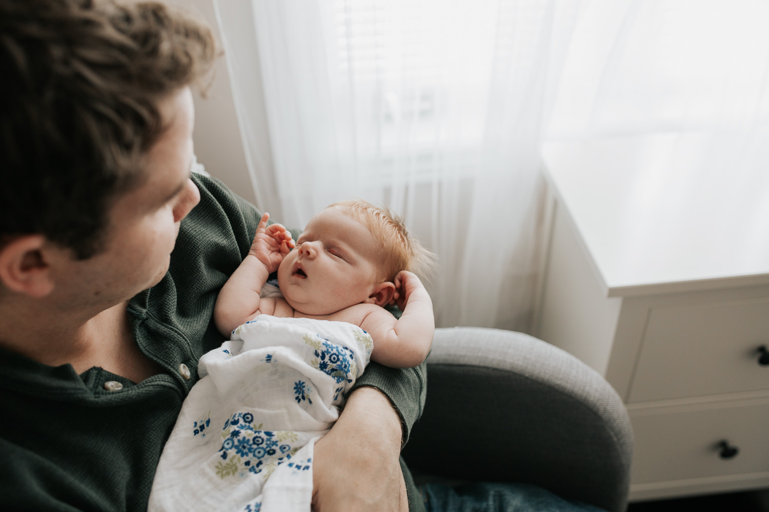 first time dad sitting in nursery chair holding sleeping 2 week old baby girl with red hair - Newmarket In-Home Photos