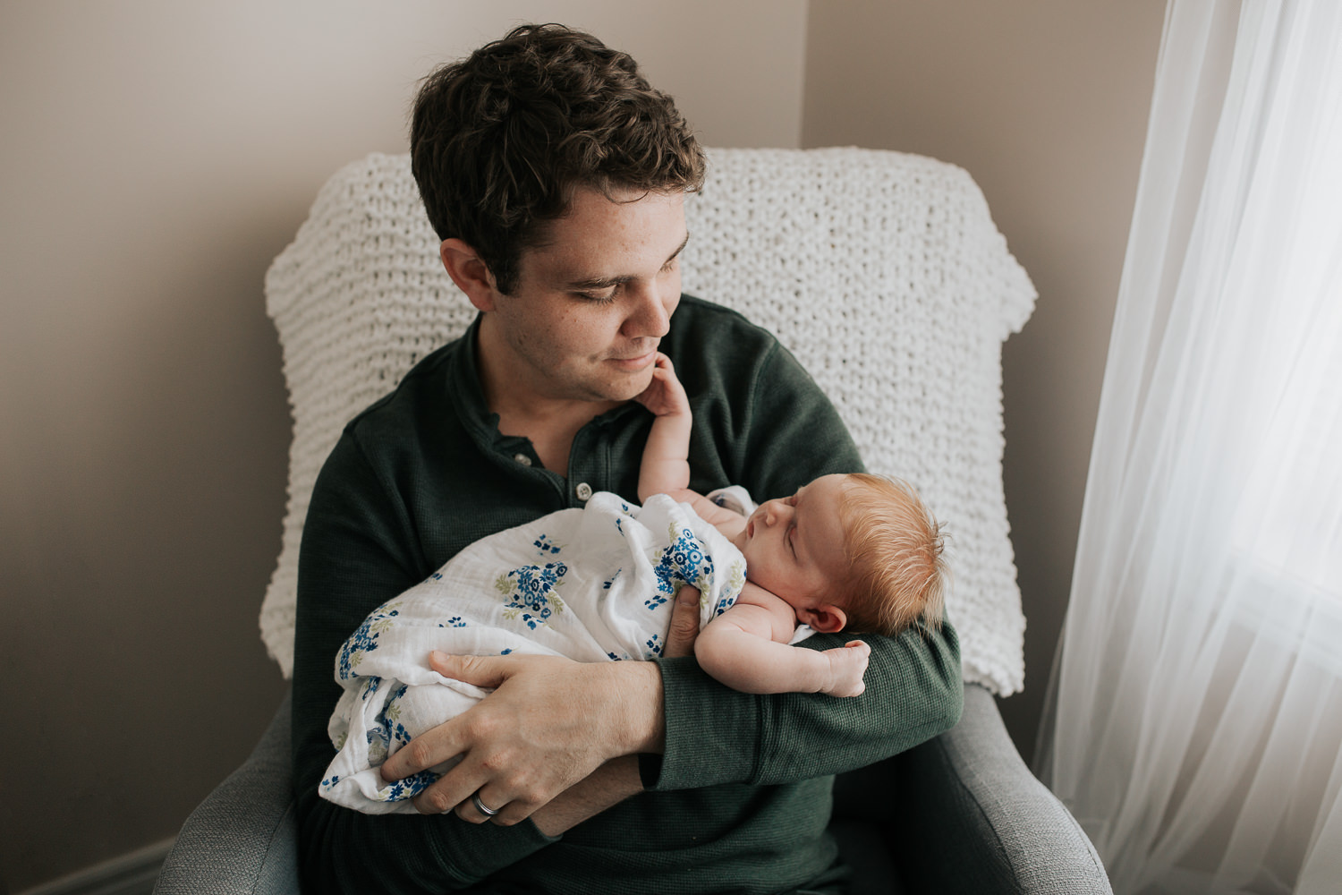 first time dad sitting in nursery chair holding sleeping 2 week old baby girl with red hair - Stouffville In-Home Photography