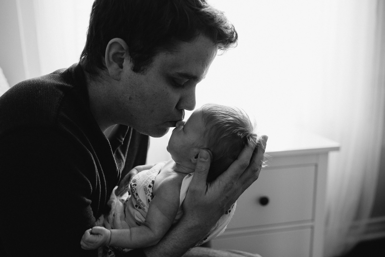 first time dad sitting in nursery chair holding sleeping 2 week old baby girl, kissing her nose - Markham In-Home Photography