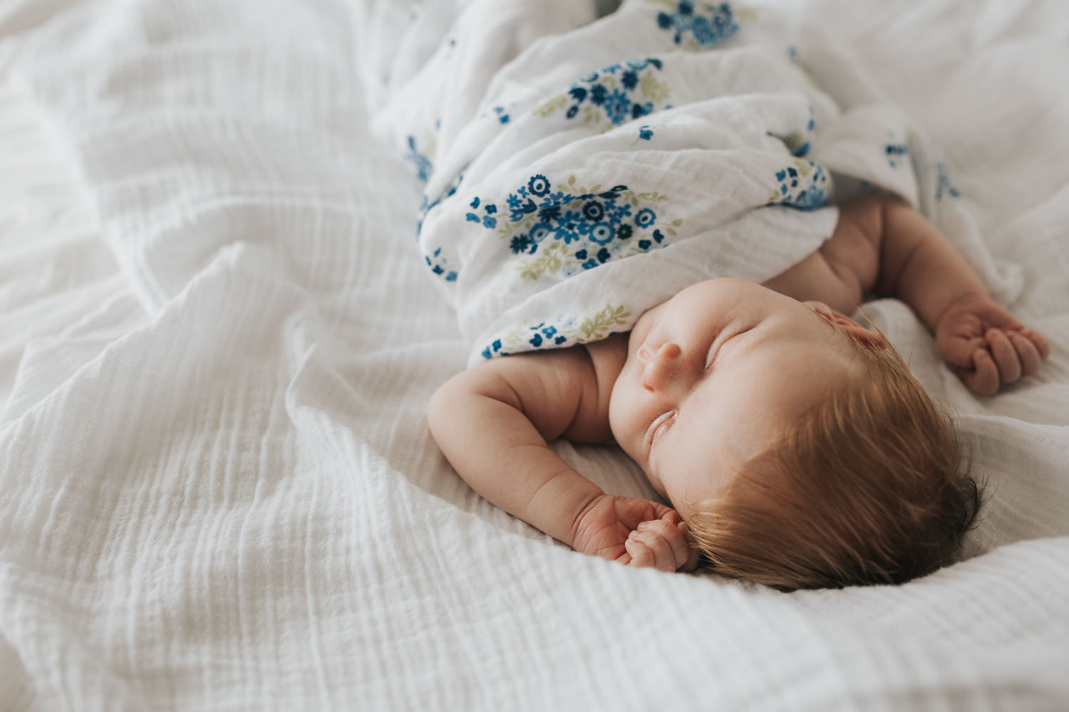 2 week old baby girl with red hair asleep on bed wrapped in blue floral swaddle - Markham Lifestyle Photography