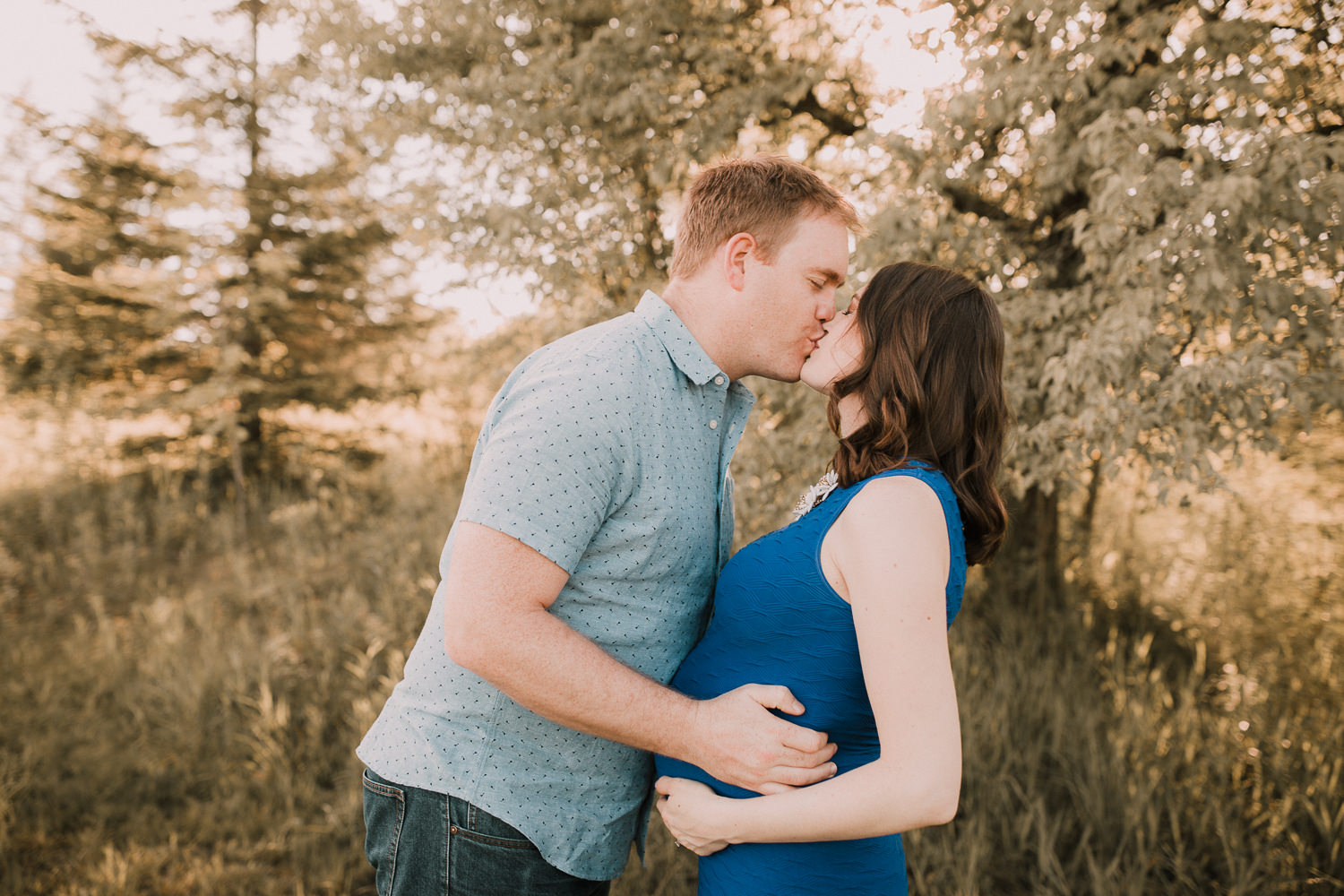 husband kisses pregnant brunette wife in blue dress while with hands on her baby bump - Stouffville Lifestyle Photography