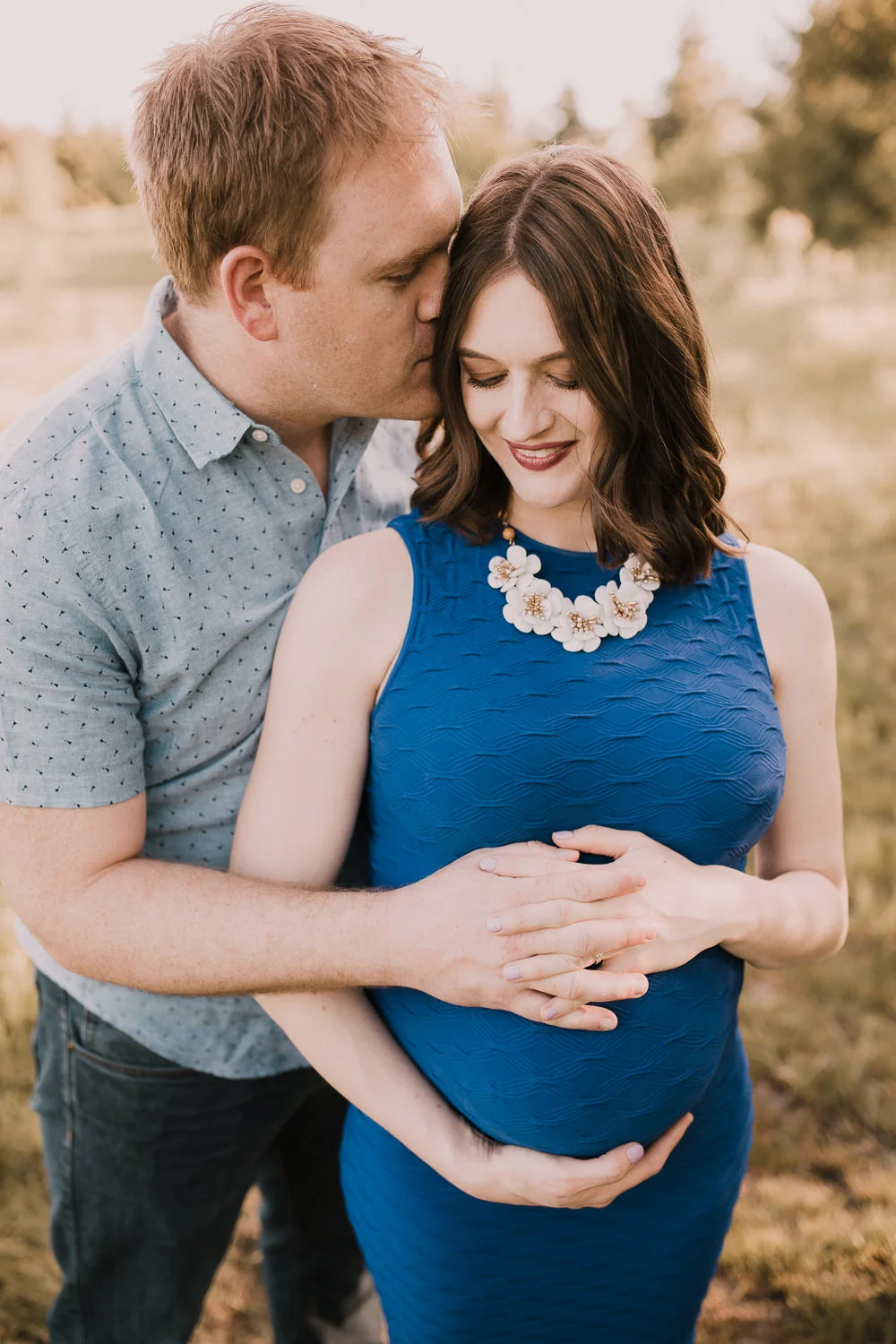 husband embraces pregnant wife in blue dress and kisses her on cheek at sunset - Barrie Lifestyle Photos