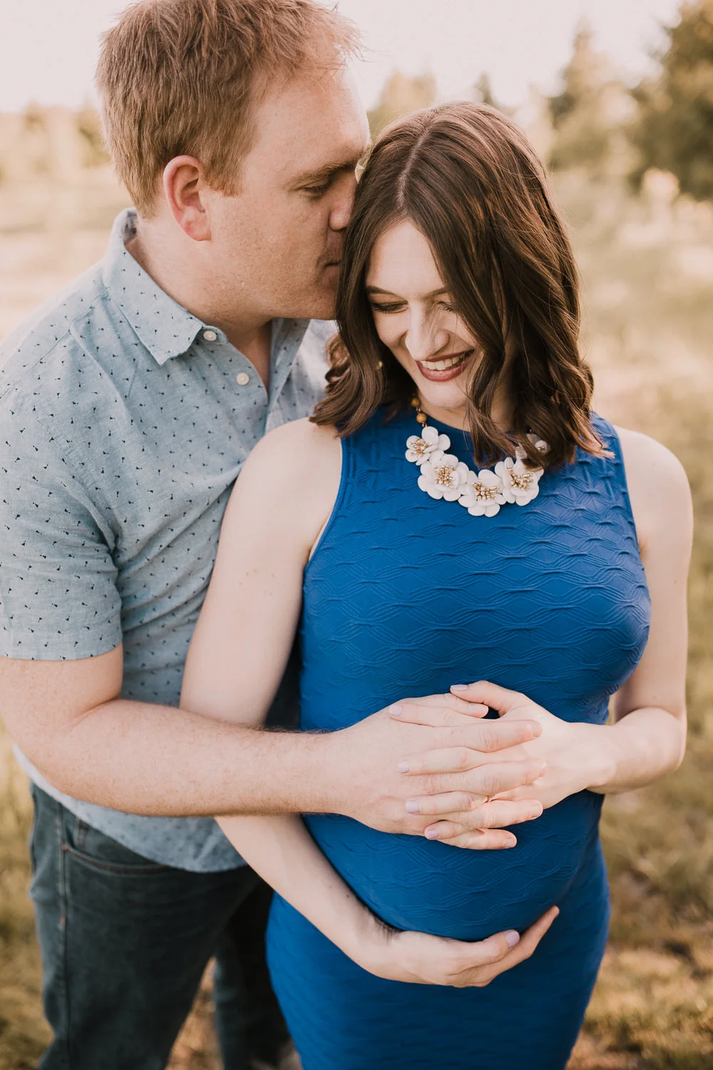 husband embraces pregnant wife in blue dress and kisses her on cheek at sunset - Newmarket Outdoor Photography