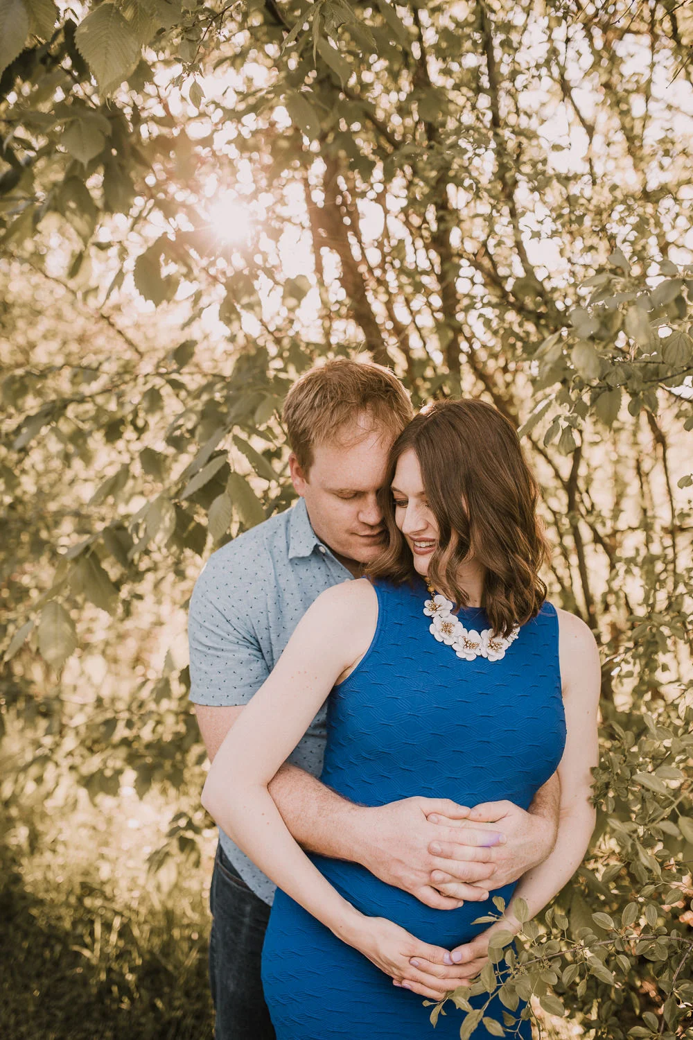 husband embraces pregnant wife in blue dress with hands on baby bump - Newmarket Outdoor Photos