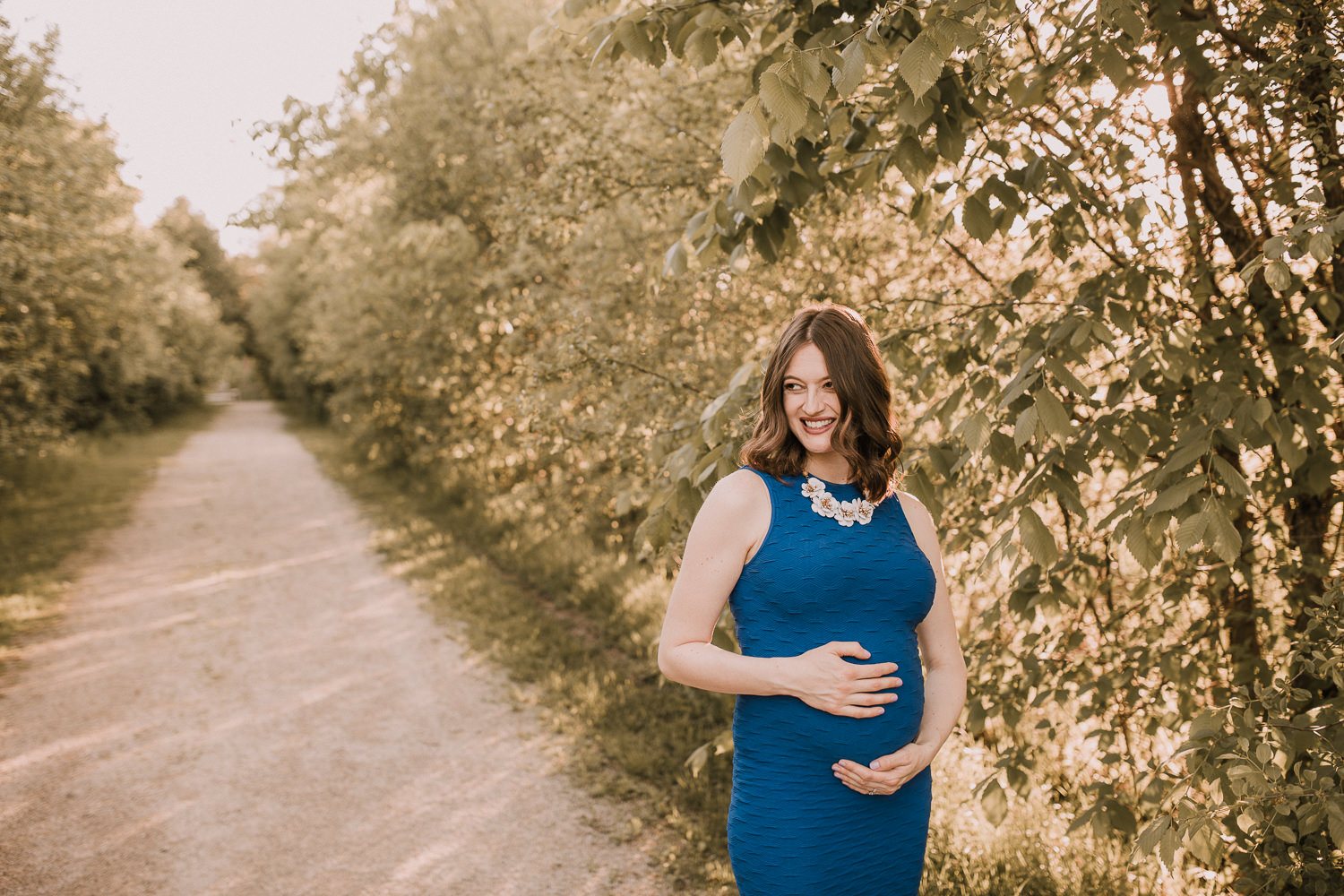 pregnant brunette mom-to-be in blue dress holding baby bump and smiling - Stouffville Outdoor Photos