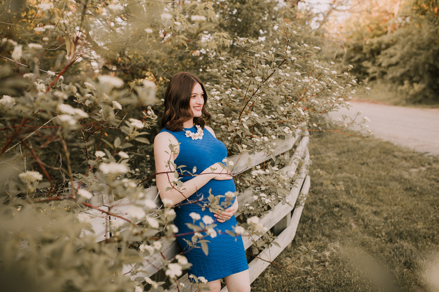 pregnant brunette mom-to-be in blue dress holding baby bump and leaning against fence - Stouffville Lifestyle Photography
