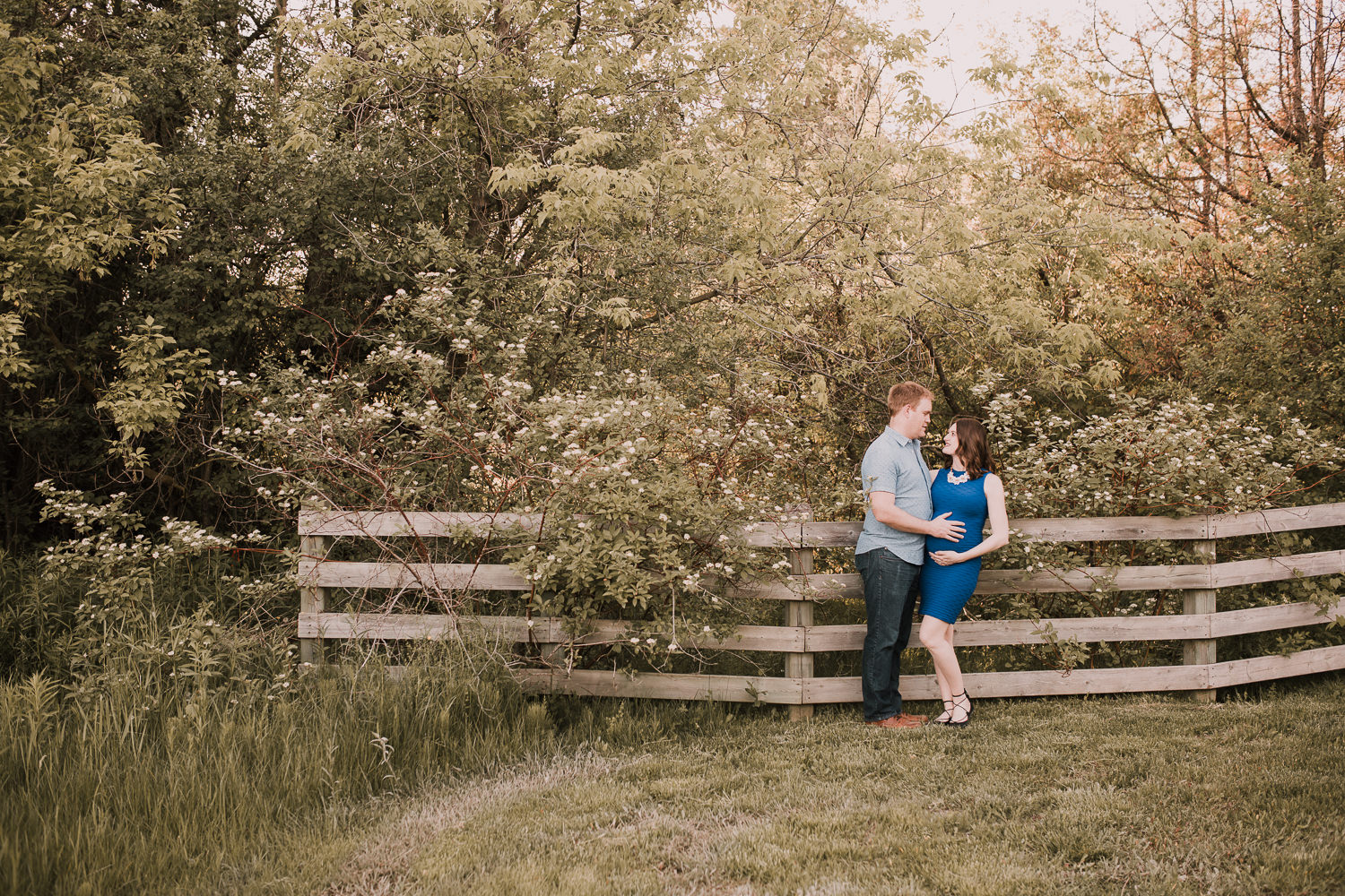 pregnant brunette mom-to-be in blue dress leaning against fence with husband, smiling at him - Newmarket Lifestyle Photos