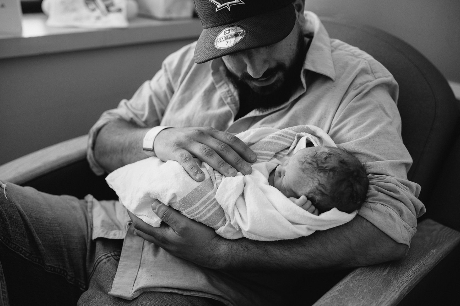 new dad sitting in hospital chair holding swaddled, sleeping 10 hour old baby boy -​​​​​​​ Newmarket In-Hospital Photography