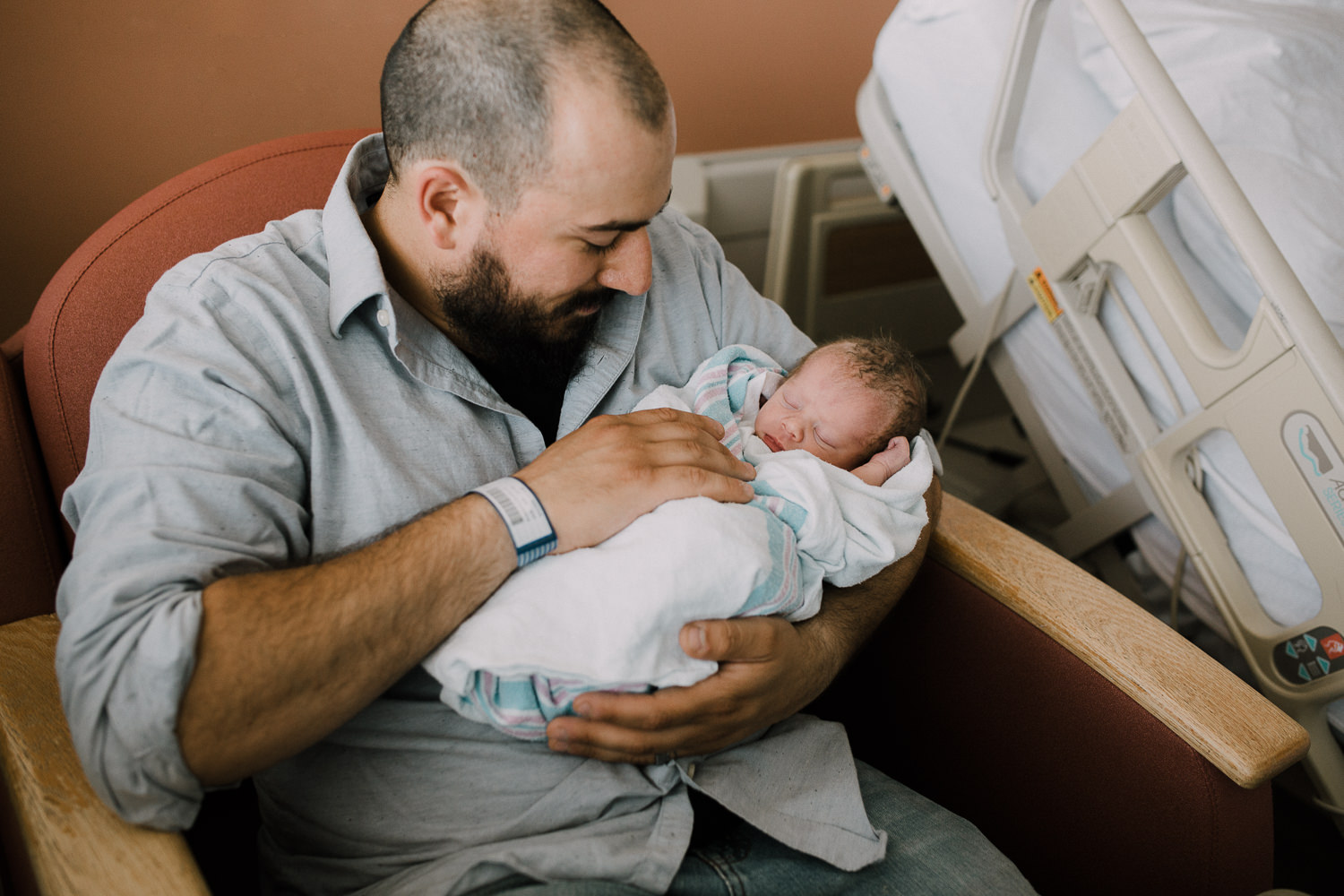 new dad sitting in hospital chair holding swaddled, sleeping 10 hour old baby boy -​​​​​​​ Barrie In-Hospital Photos