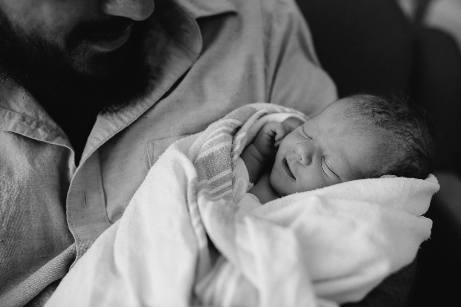 new dad sitting in hospital chair holding sleeping, smiling 10 hour old baby boy -​​​​​​​ Markham In-Hospital Photography