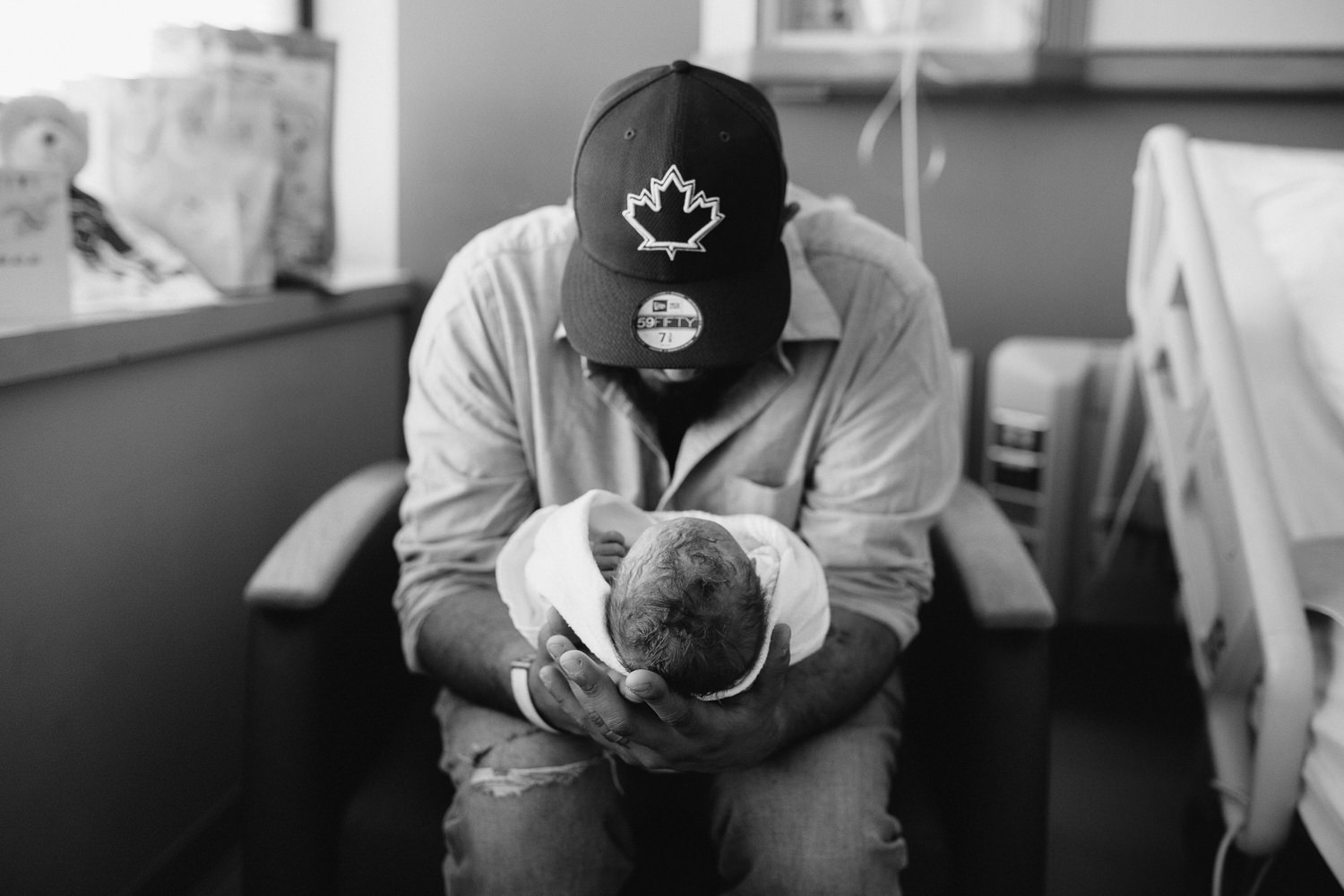 new dad sitting in hospital chair holding swaddled, sleeping 10 hour old baby boy -​​​​​​​ Markham Fresh 48 Photography