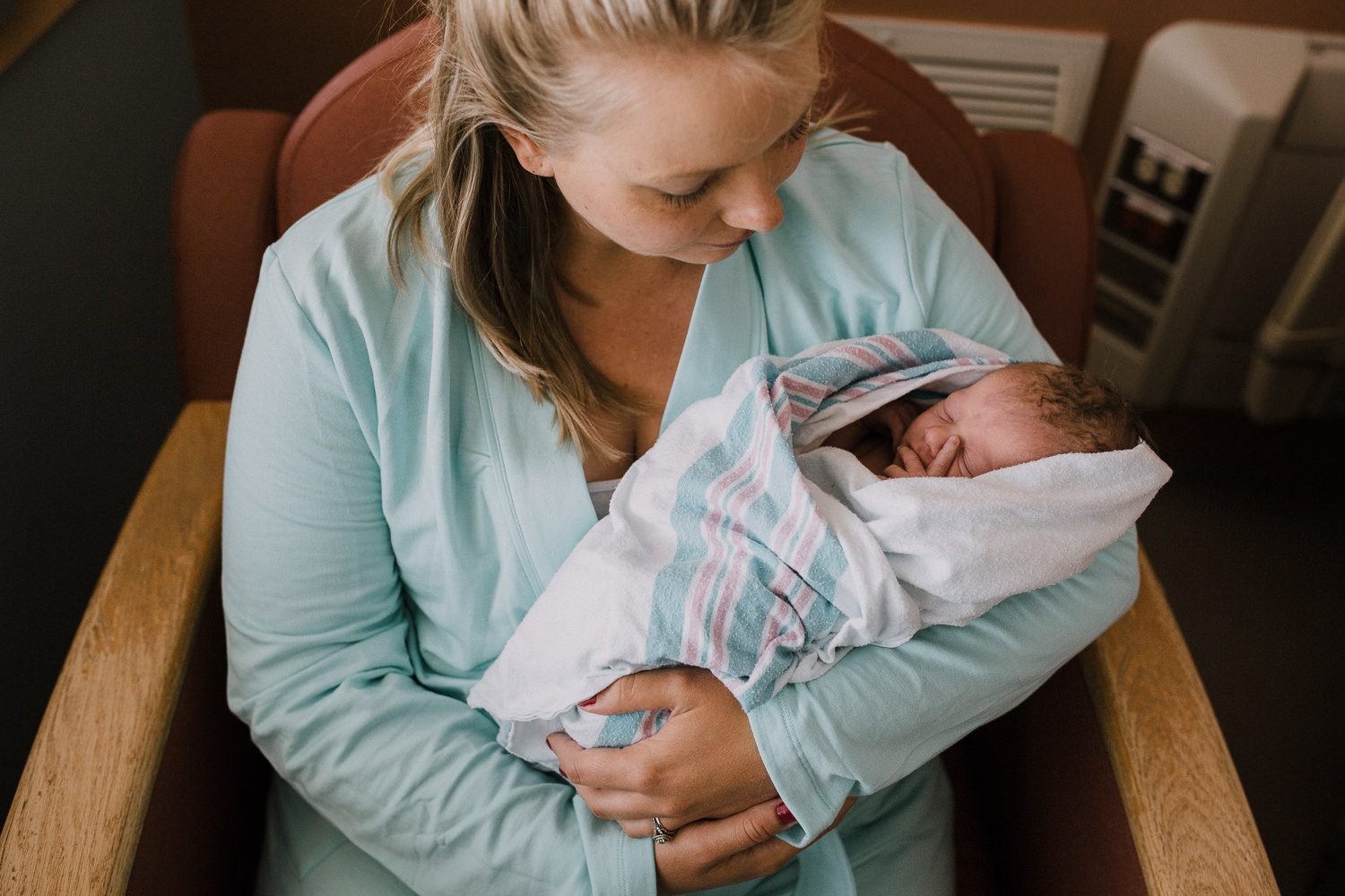 new mother sitting in hospital chair holding swaddled 10 hour old baby boy -​​​​​​​ Newmarket In-Hospital Photos