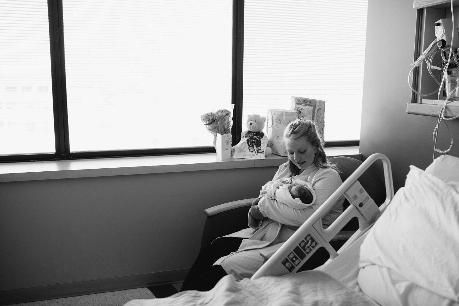 new mother sitting in hospital chair holding swaddled 10 hour old baby boy -​​​​​​​ Newmarket In-Hospital Photography