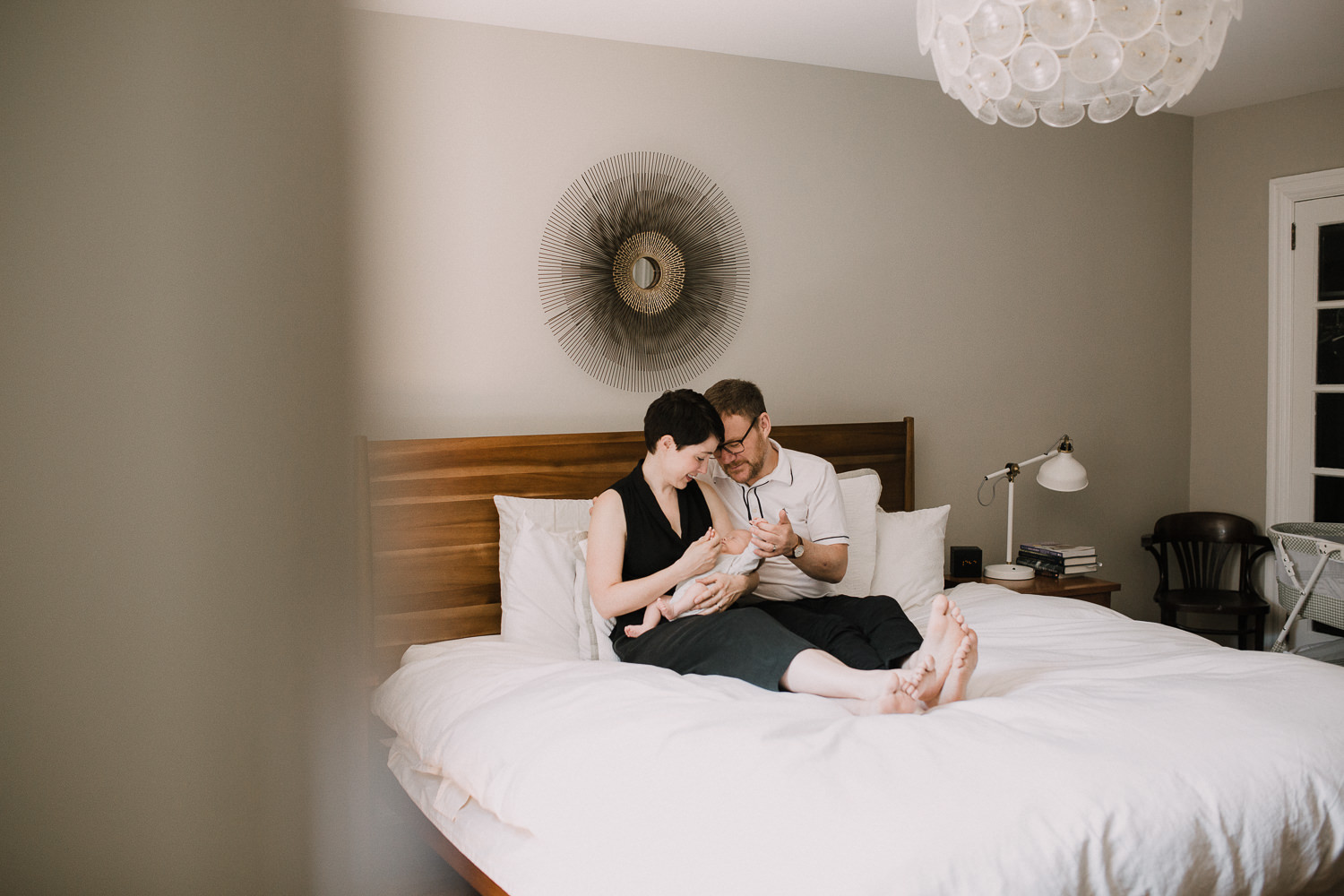 new parents sitting in master bed holding and looking at 2 week old sleeping baby boy - Newmarket Lifestyle Photography