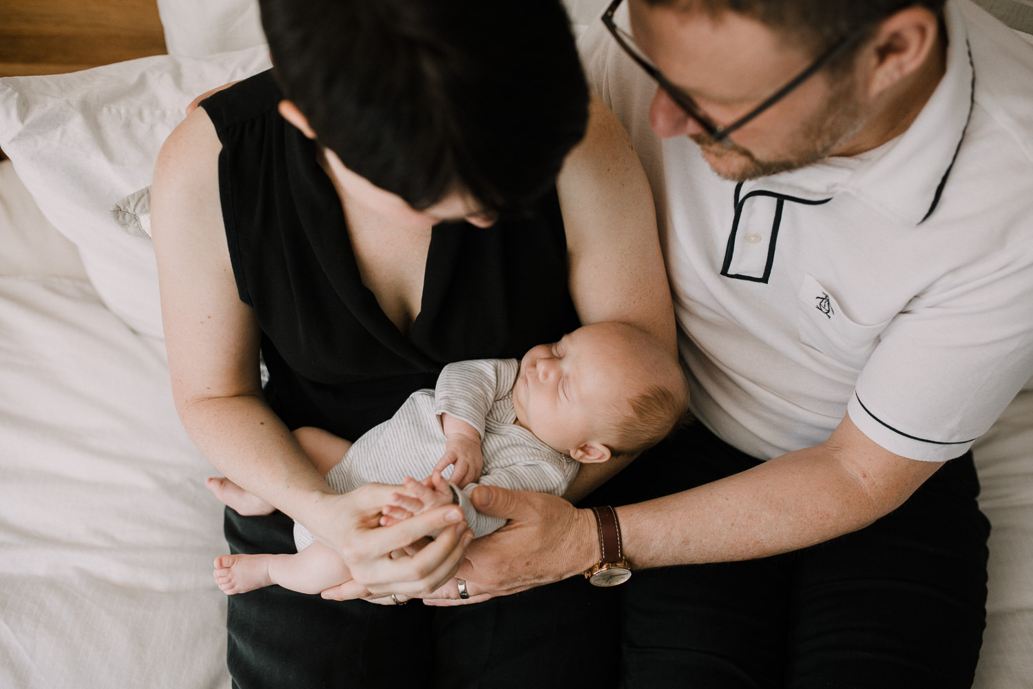 new parents sitting in master bed holding and looking at 2 week old sleeping baby boy - Markham Lifestyle Photography