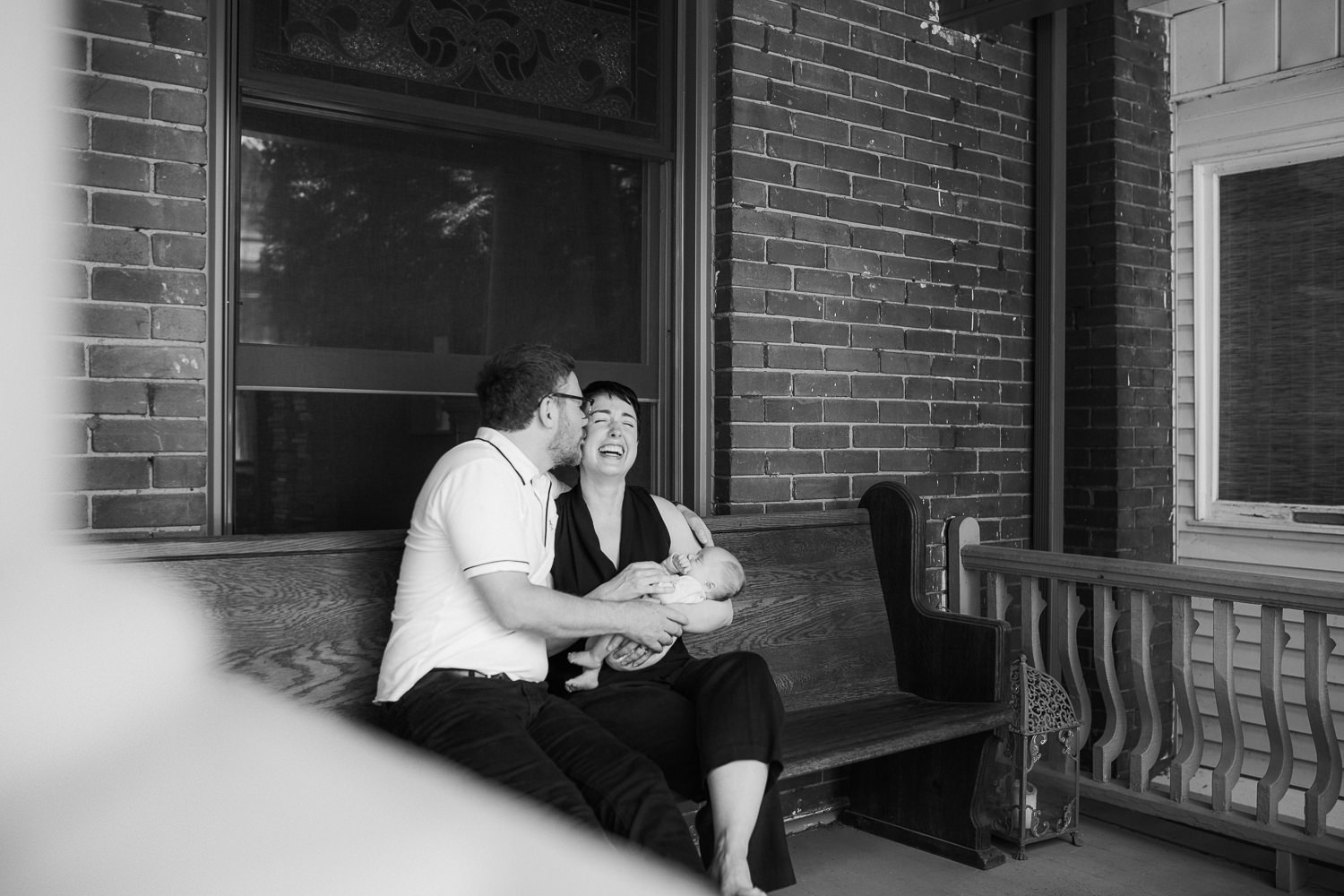 new parents sitting on front porch holding sleeping 2 week old baby boy, dad kissing mom's cheek - Barrie In-Home Photos