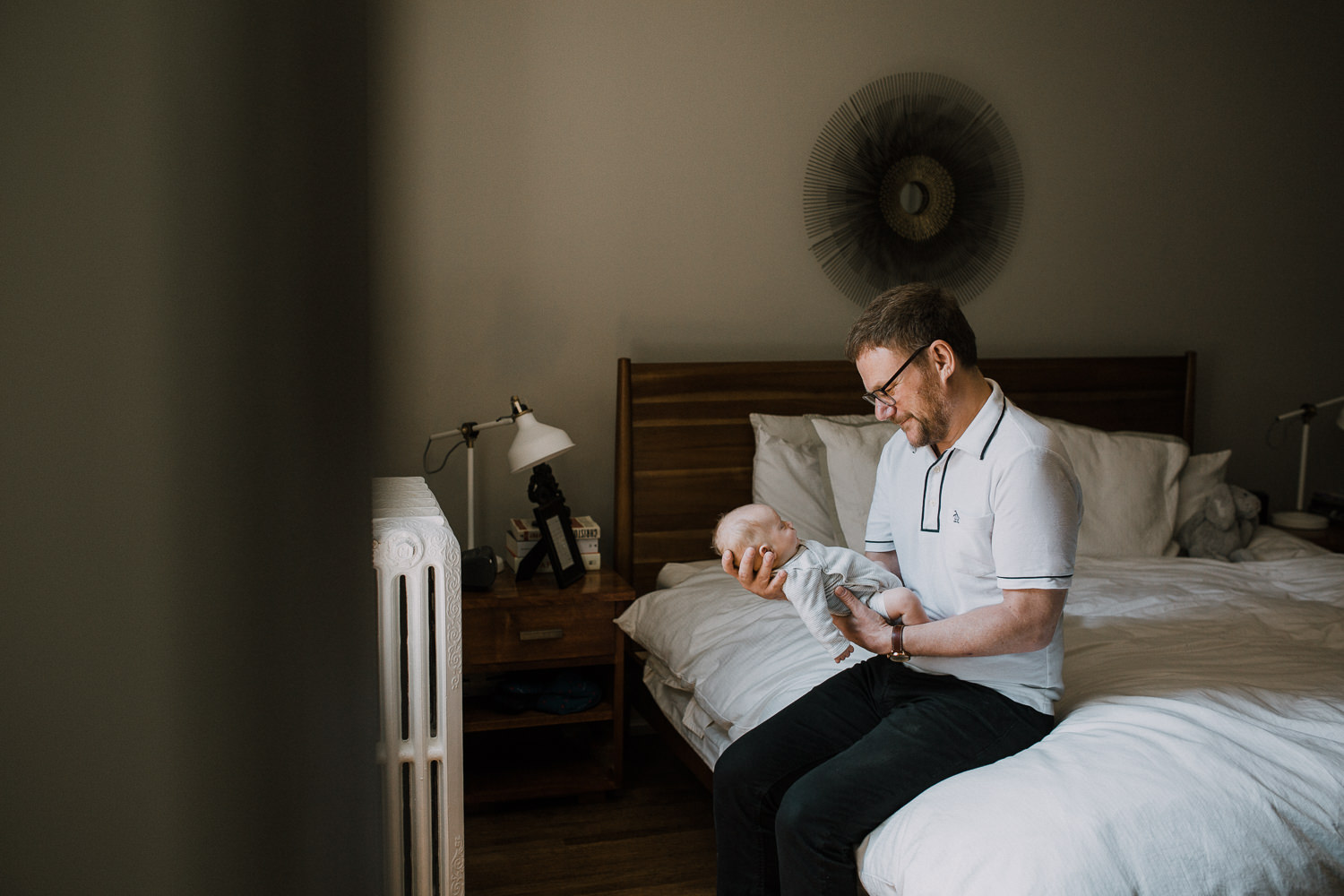 first time father sitting on master bed holding and looking at 2 week old baby boy - Stouffville In-Home Photography