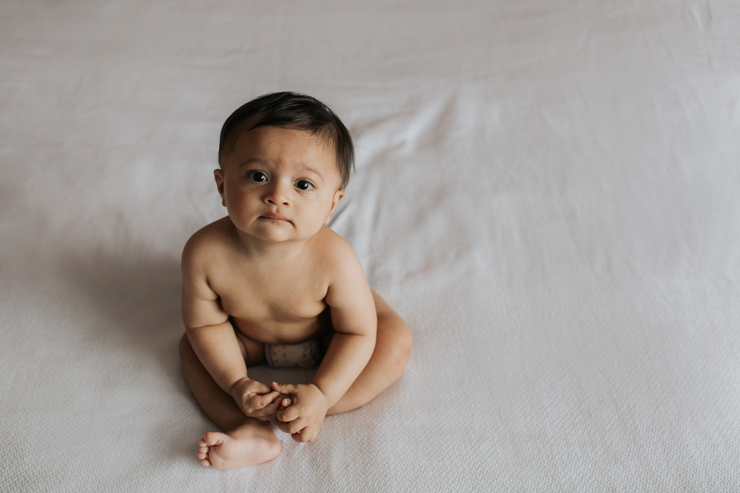 8 month old baby boy in diaper sitting on bed looking at camera - Markham Lifestyle Photography