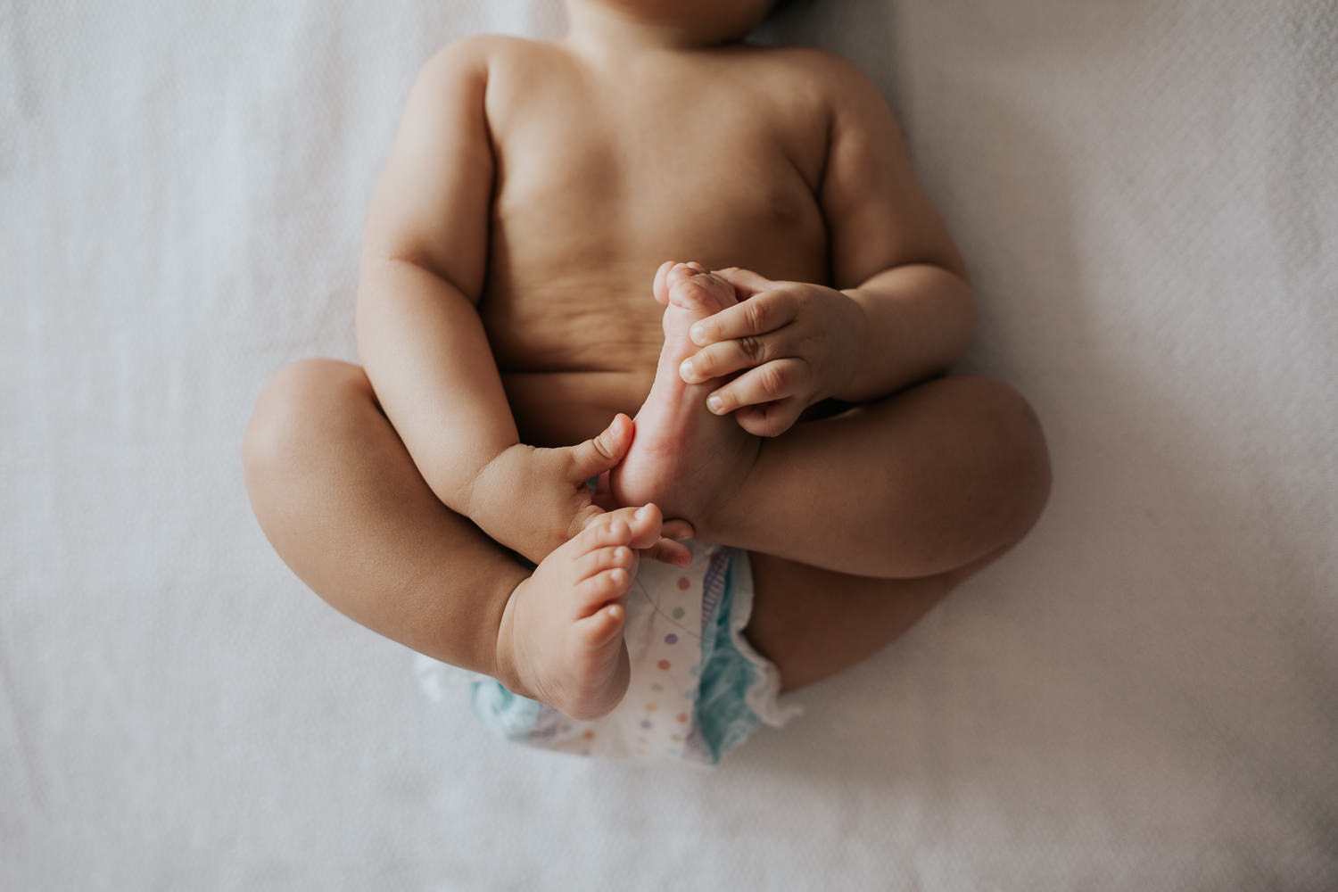 8 month old baby boy in diaper lying on bed, playing with his feet - Newmarket In-Home Photos