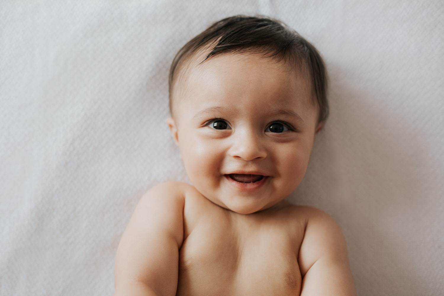 8 month old baby boy with brown hair and eyes in diaper lying on bed smiling looking at camera - Markham In-Home Photography