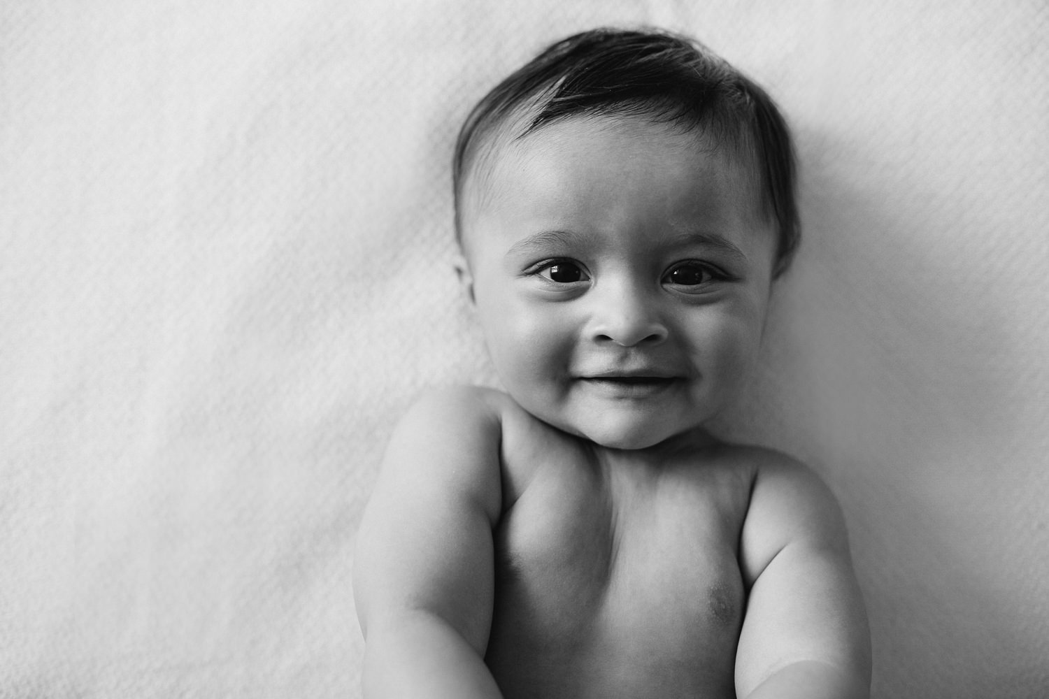 8 month old baby boy with brown hair and eyes in diaper lying on bed smiling looking at camera - Barrie In-Home Photography