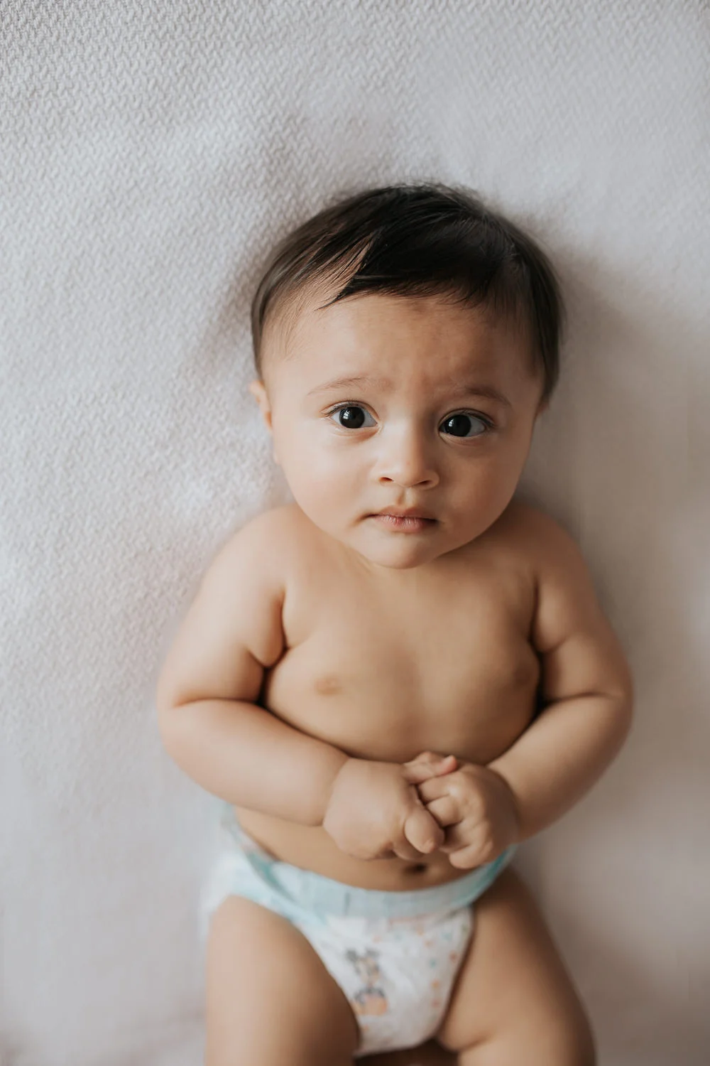 8 month old baby boy with brown hair and eyes in diaper lying on bed, hands folded - Markham In-Home Photos