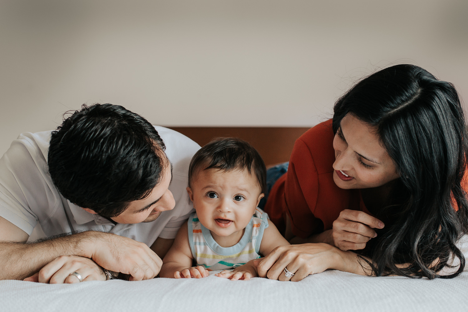 parents lying on bed with 8 month old baby boy between them doing tummy time and looking at camera - Newmarket Lifestyle Photos