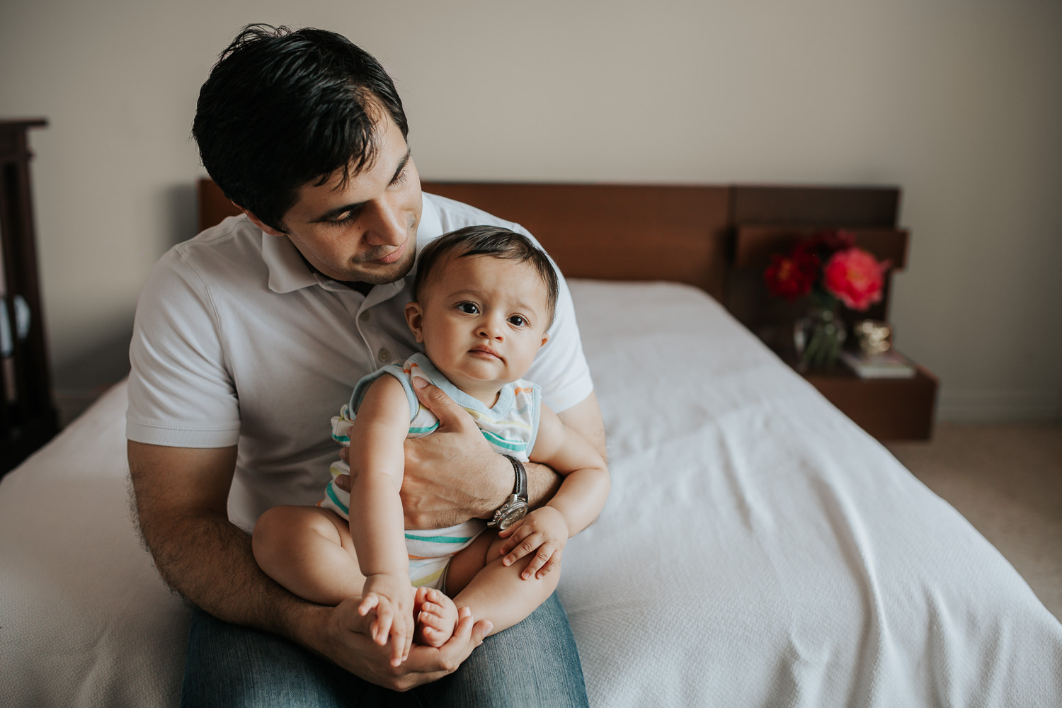 father sitting on bed with 8 month old baby boy in his lap, smiling at son who looks on - Stouffville In-Home Photography