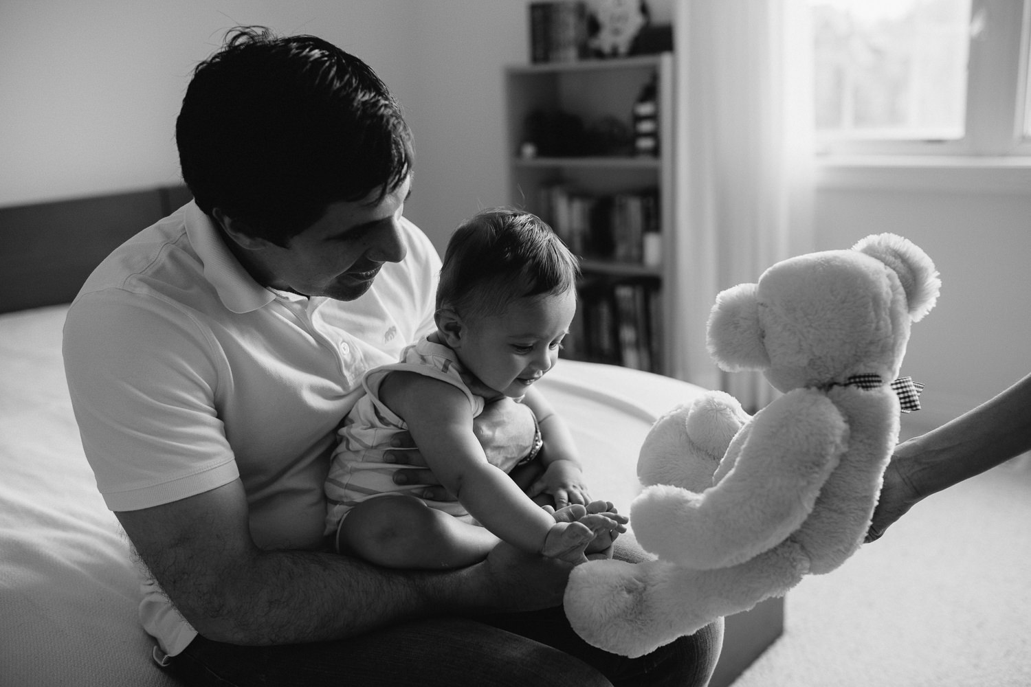 father sitting on bed with 8 month old baby boy in his lap, baby reaching for teddy bear offered by mother - Newmarket In-Home Photos