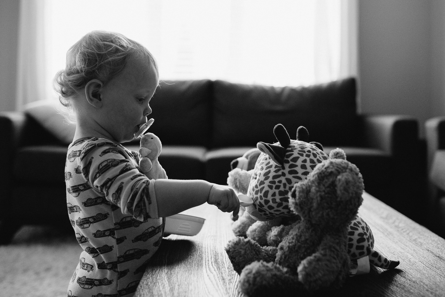 blonde 18 month old boy pretending to feed stuffed animals lined up on table - Newmarket Documentary Photography