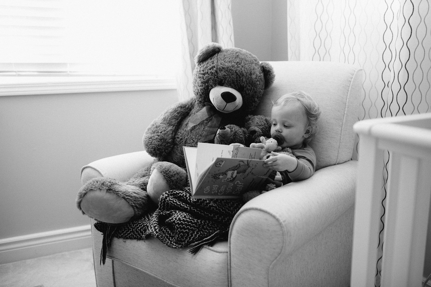 blonde 18 month boy sitting in glider with giant stuffed teddy bear reading a book - Barrie Documentary Photography