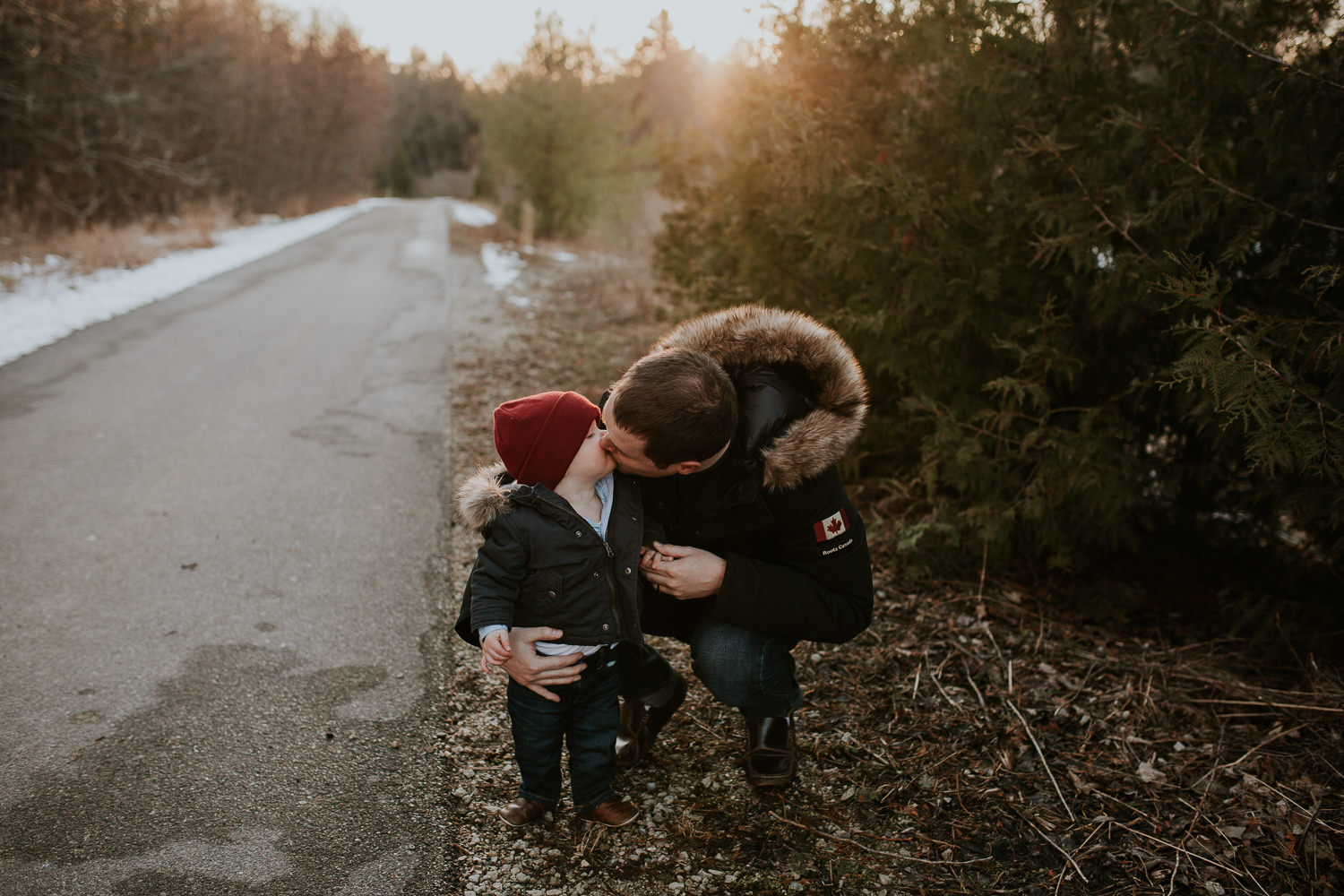 18 month old boy in red hat and parka kissing his dad on a wooded trail - Newmarket Family Photographs