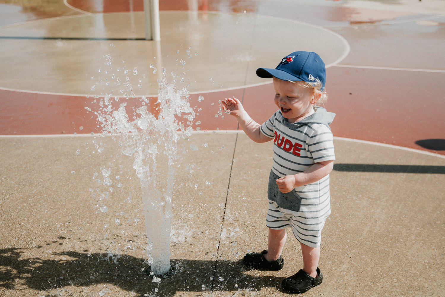 toddler boy in baseball cap laughing while playing in splash pad - Markham Documentary Photographer