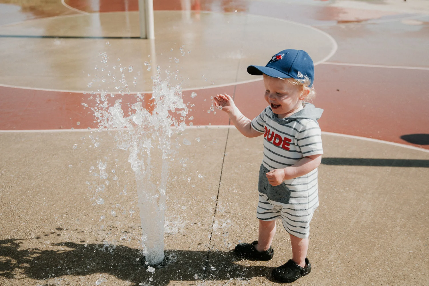 toddler boy in baseball cap laughing while playing in splash pad - Markham Documentary Photographer