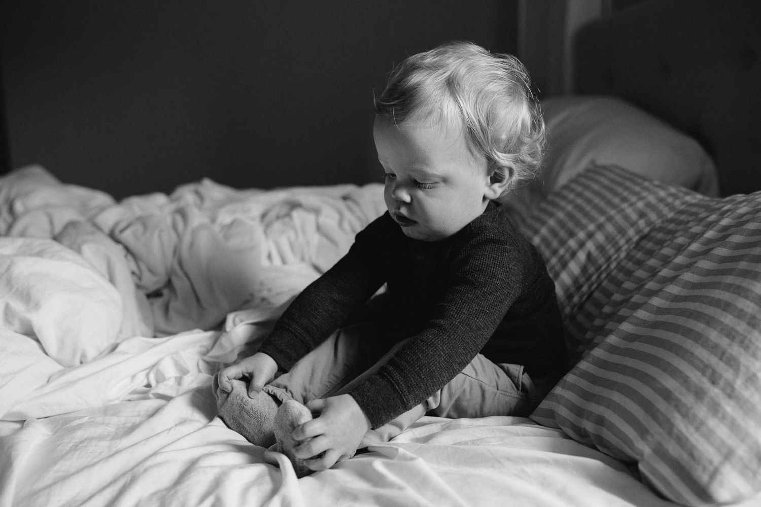 blonde 18 month toddler sitting on bed holding his feet - Markham Documentary Photographs