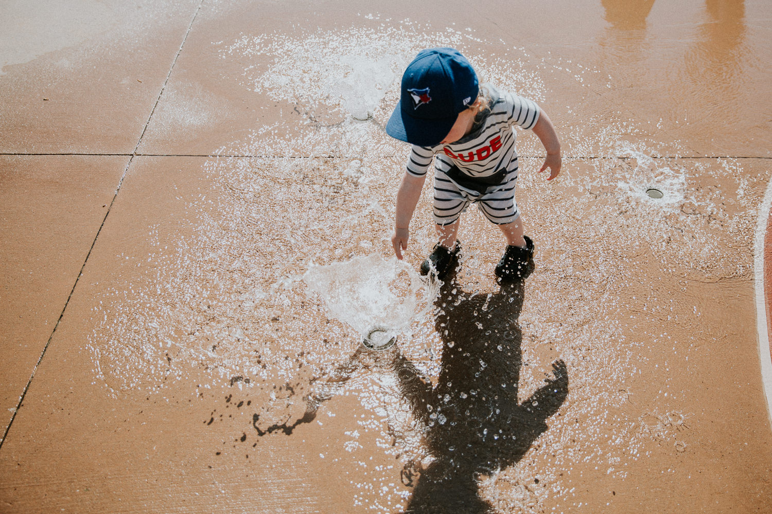 toddler boy in baseball cap playing at water splash pad -  Barrie Documentary Photographer