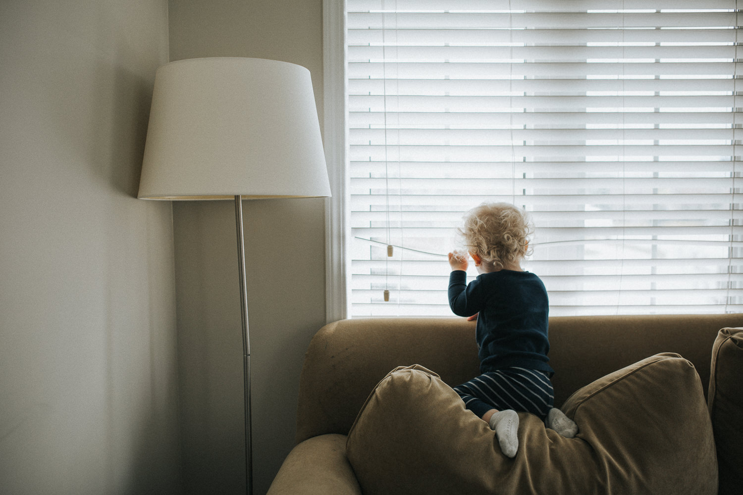 blonde toddler with curly hair sitting on top of couch pulling back blinds to look out window - Barrie Family Memories