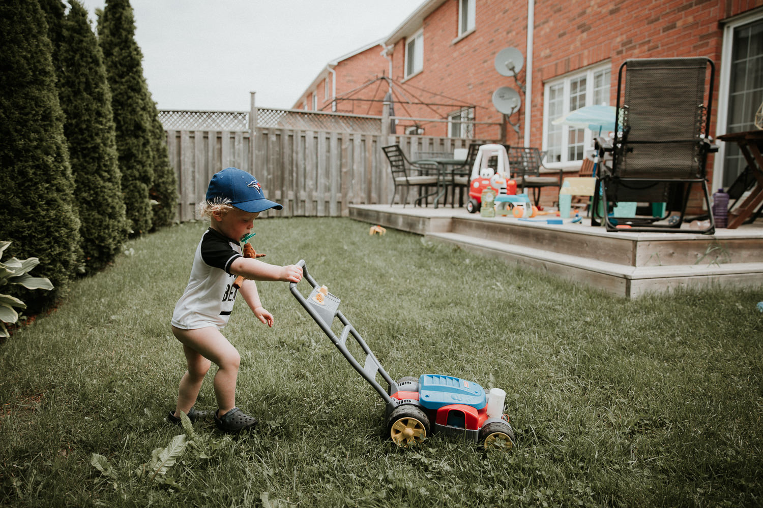 20 month old boy pushing toy lawn mower across backyard - Stouffville Documentary Photography