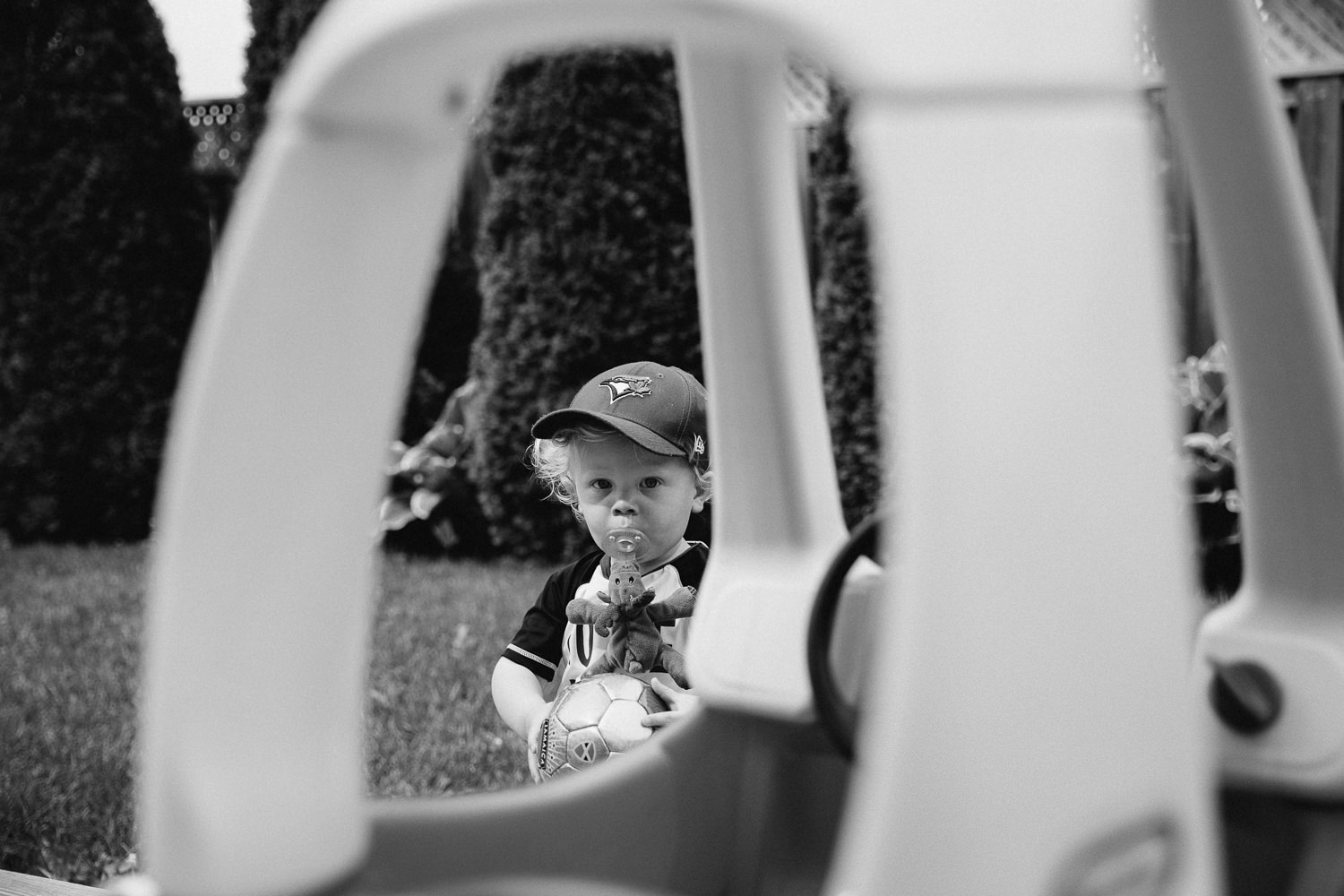 toddler boy in baseball cap with soother in mouth playing in backyard, looking at camera - Markham Documentary Photography