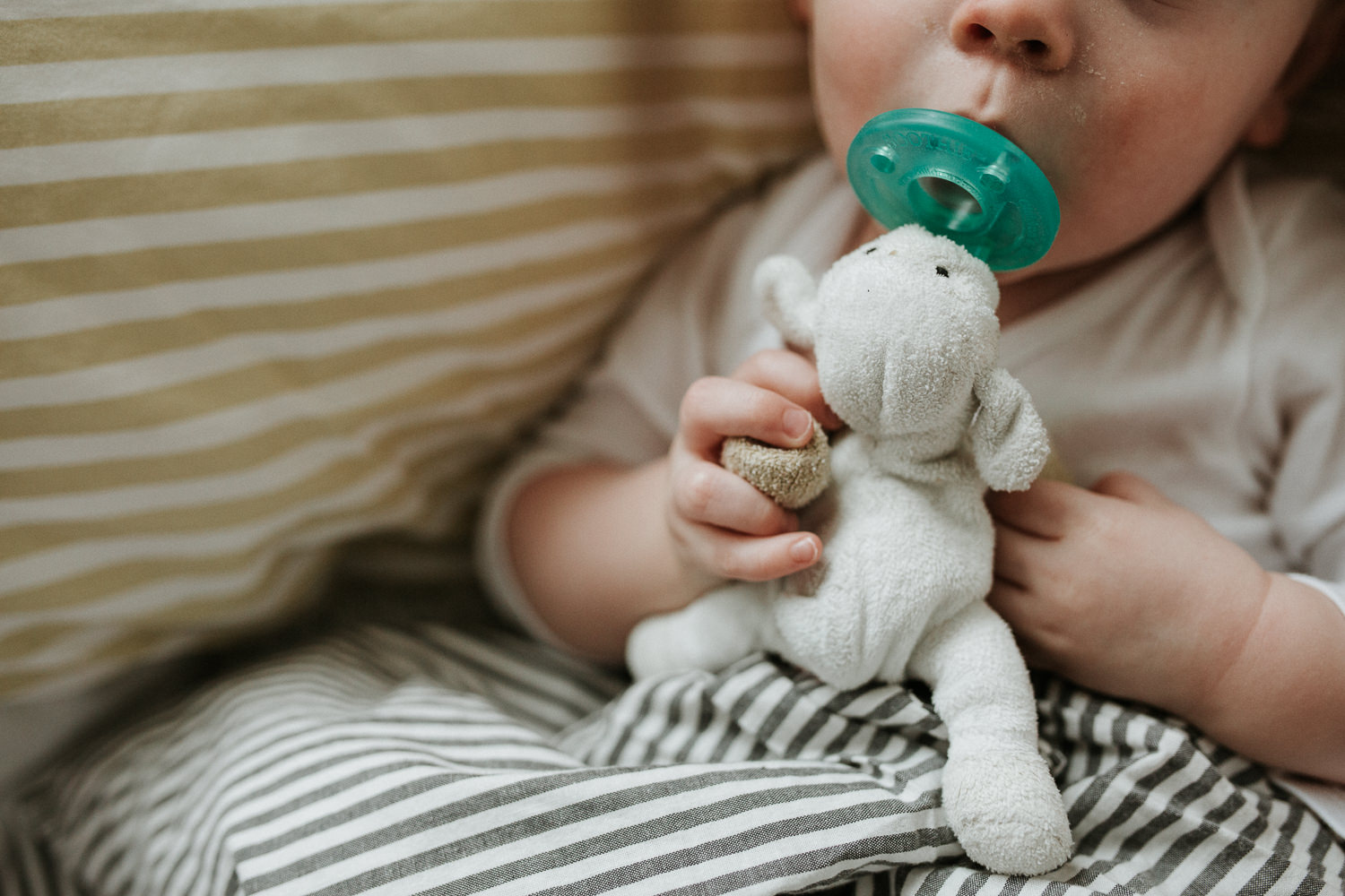 toddler lying in bed with striped linens, close up of him holding wubbanub soother - Markham Family Photography