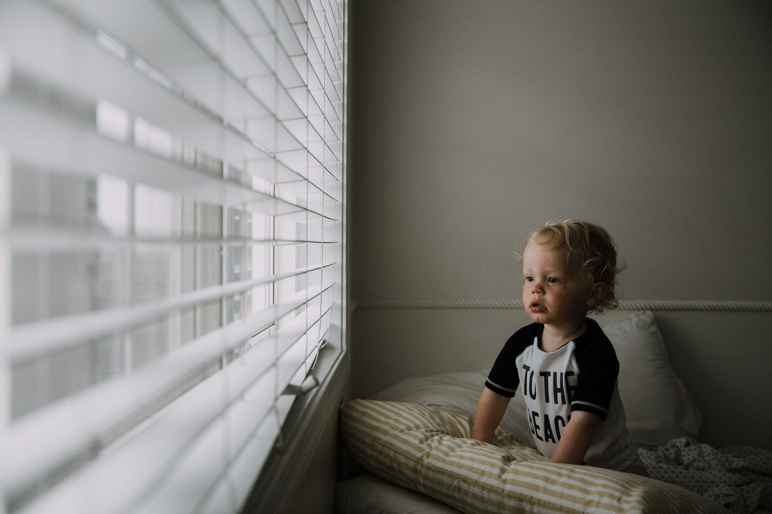 toddler boy with curly blonde hair sitting on bed looking out window at a thunderstorm - Markham Family Photography