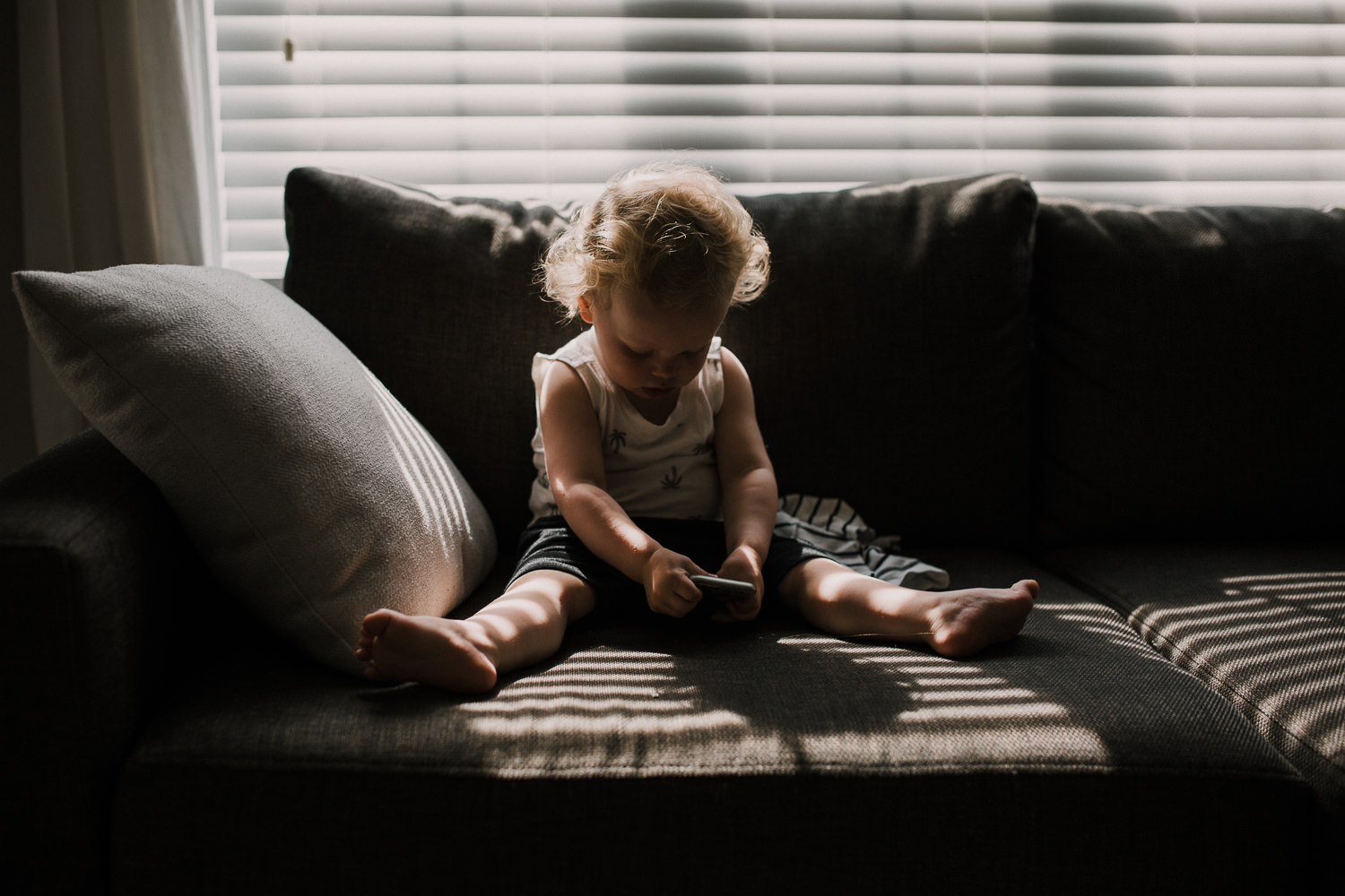 18 month old boy with curly hair sitting in harsh light on couch looking at phone - Stouffville Documentary Photographer