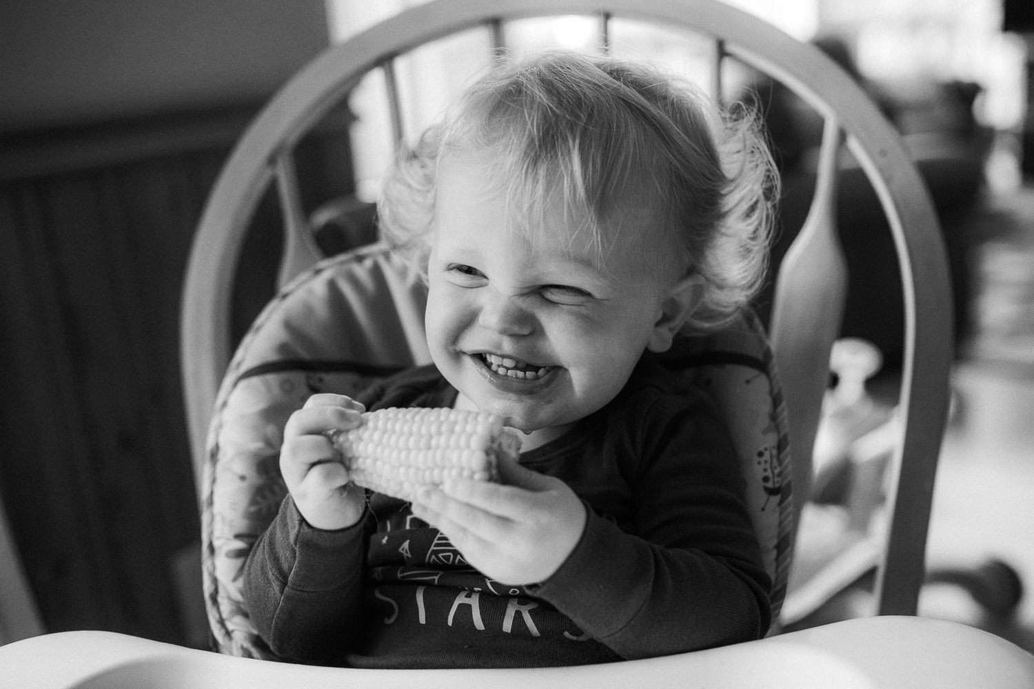 blonde toddler boy smiling in his high chair while eating corn on the cob - Stouffville Documentary Photographer