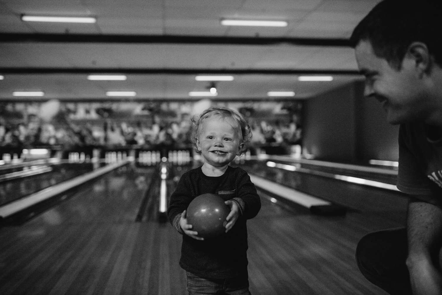 18 month old boy standing in bowling lane holding ball and smiling, dad off to side - Stouffville Family Memories