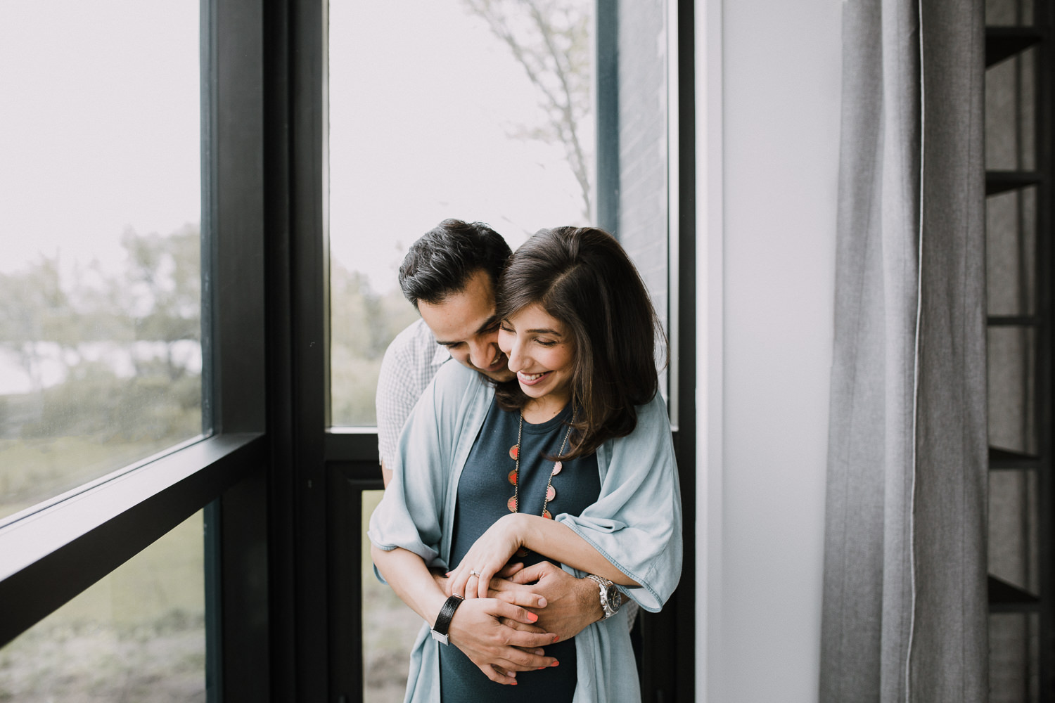 husband embraces pregnant wife from behind, standing next to window - Newmarket Lifestyle Photography