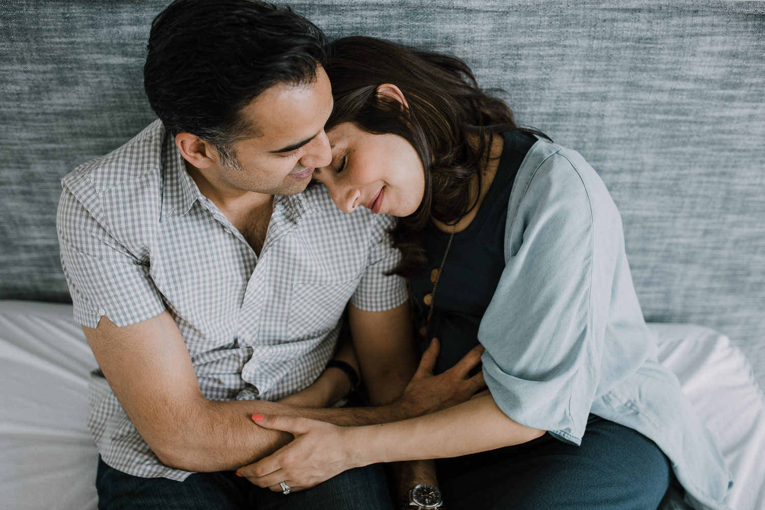 couple sitting on bed, wife resting head on husband's shoulder while he rests his hand on her pregnant belly - Markham In-Home Photography