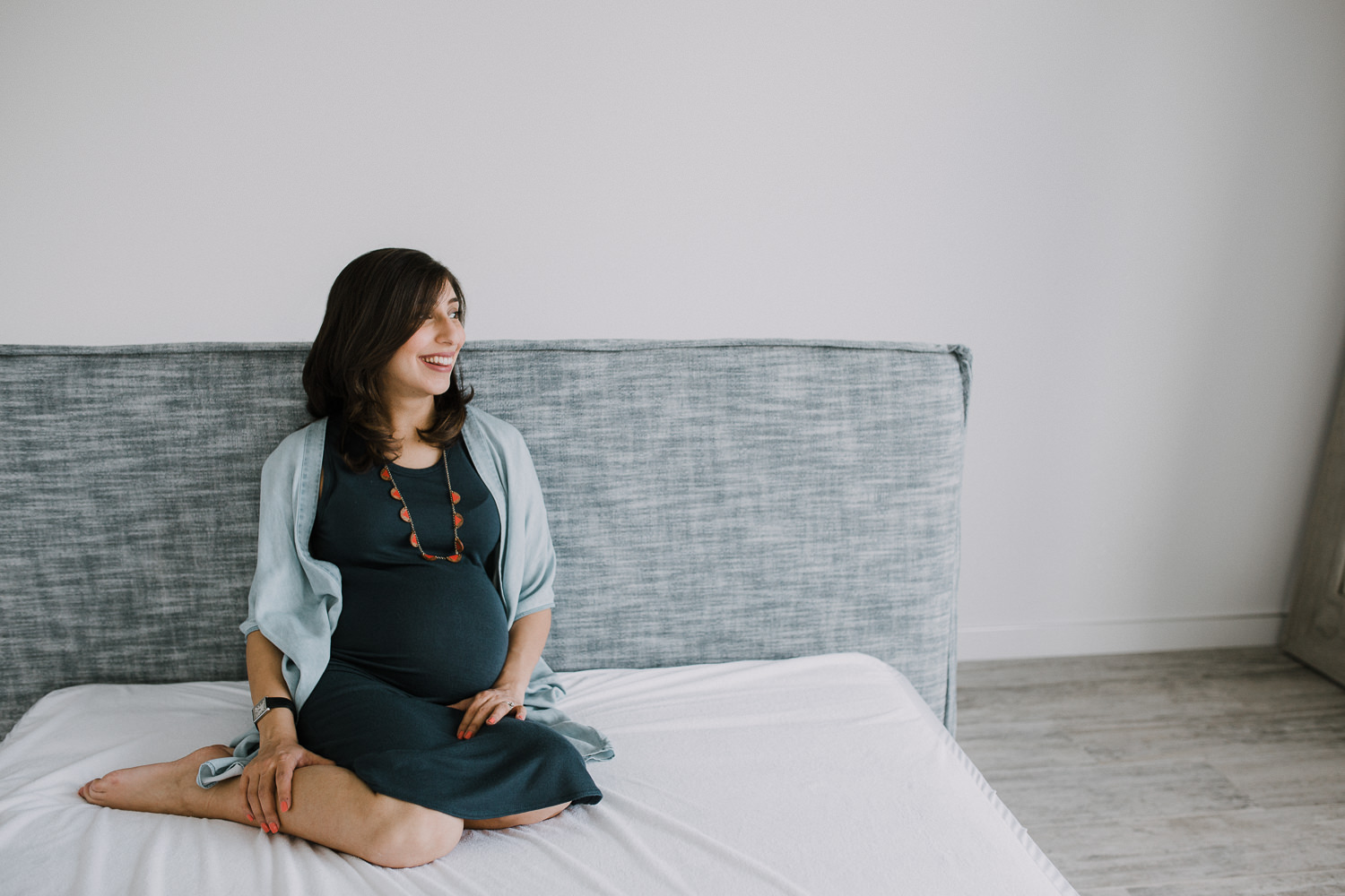 pregnant woman sitting on bed, smiling and looking out window - Barrie In-Home Photography