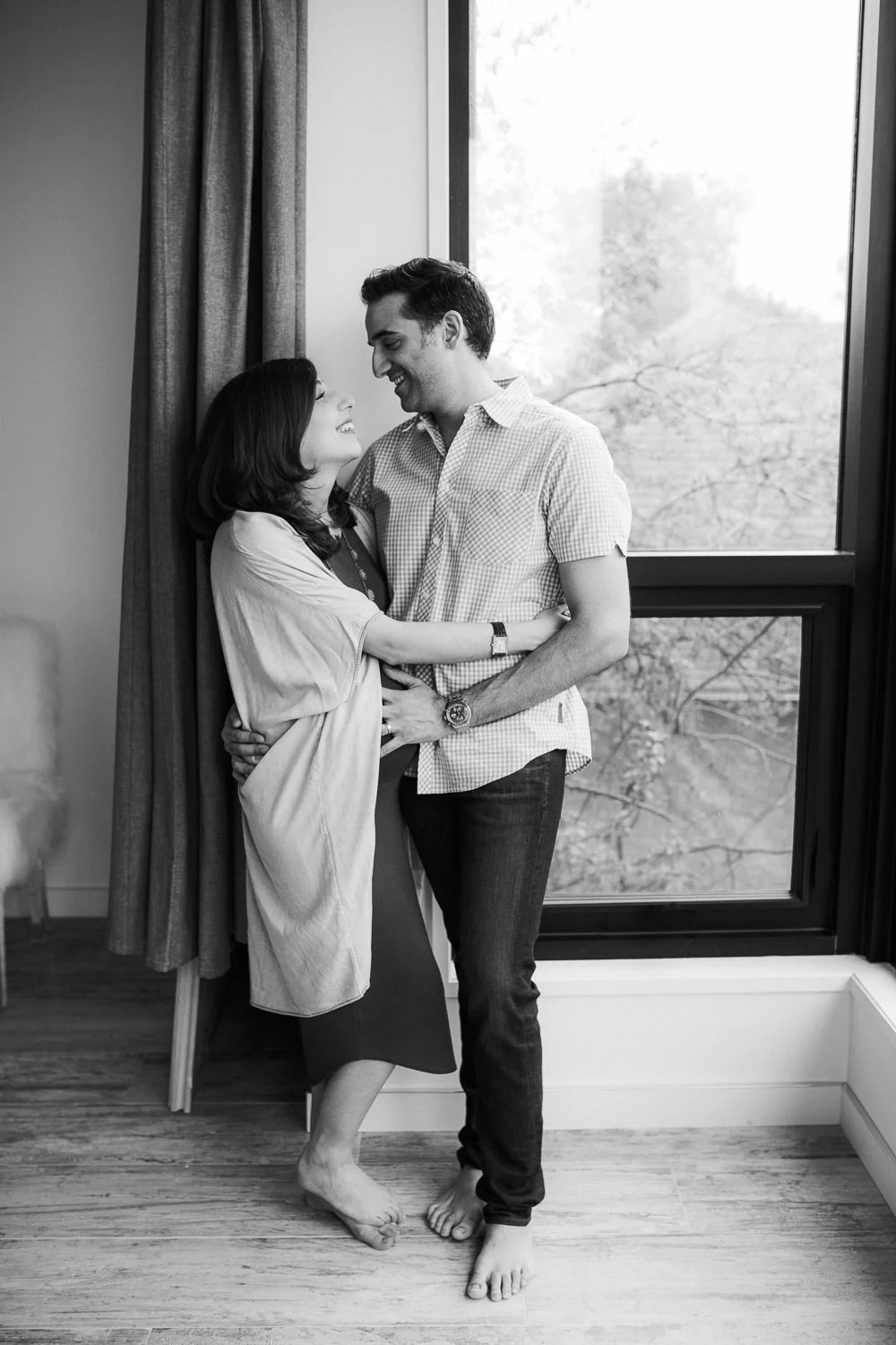 husband and pregnant wife embracing and smiling at one another, standing next to window - Barrie In-Home Photos
